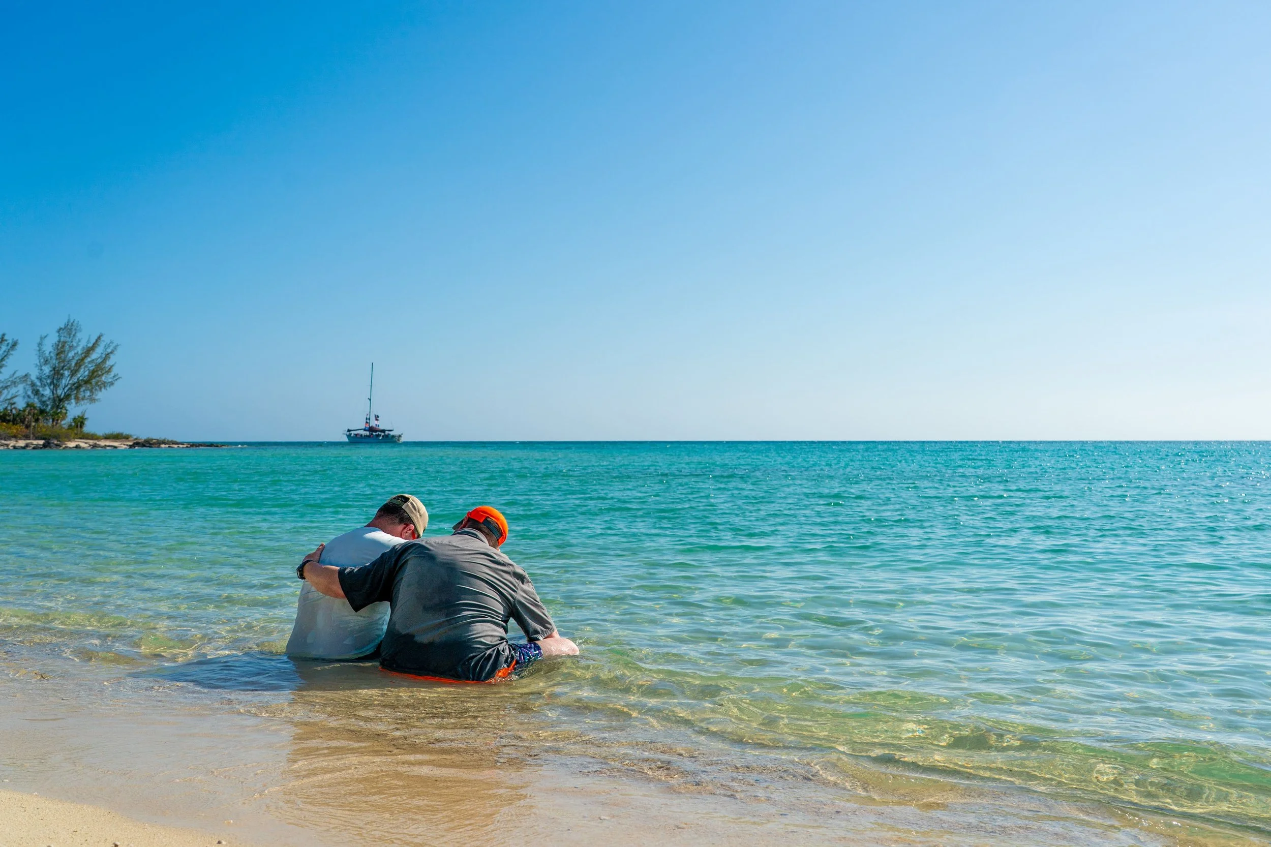 Two men sitting on a sandy beach near the water's edge, with one in a white shirt and the other in a gray shirt and orange cap, taking a break and looking out at the ocean, with a sailboat in the distance on a clear, sunny day.