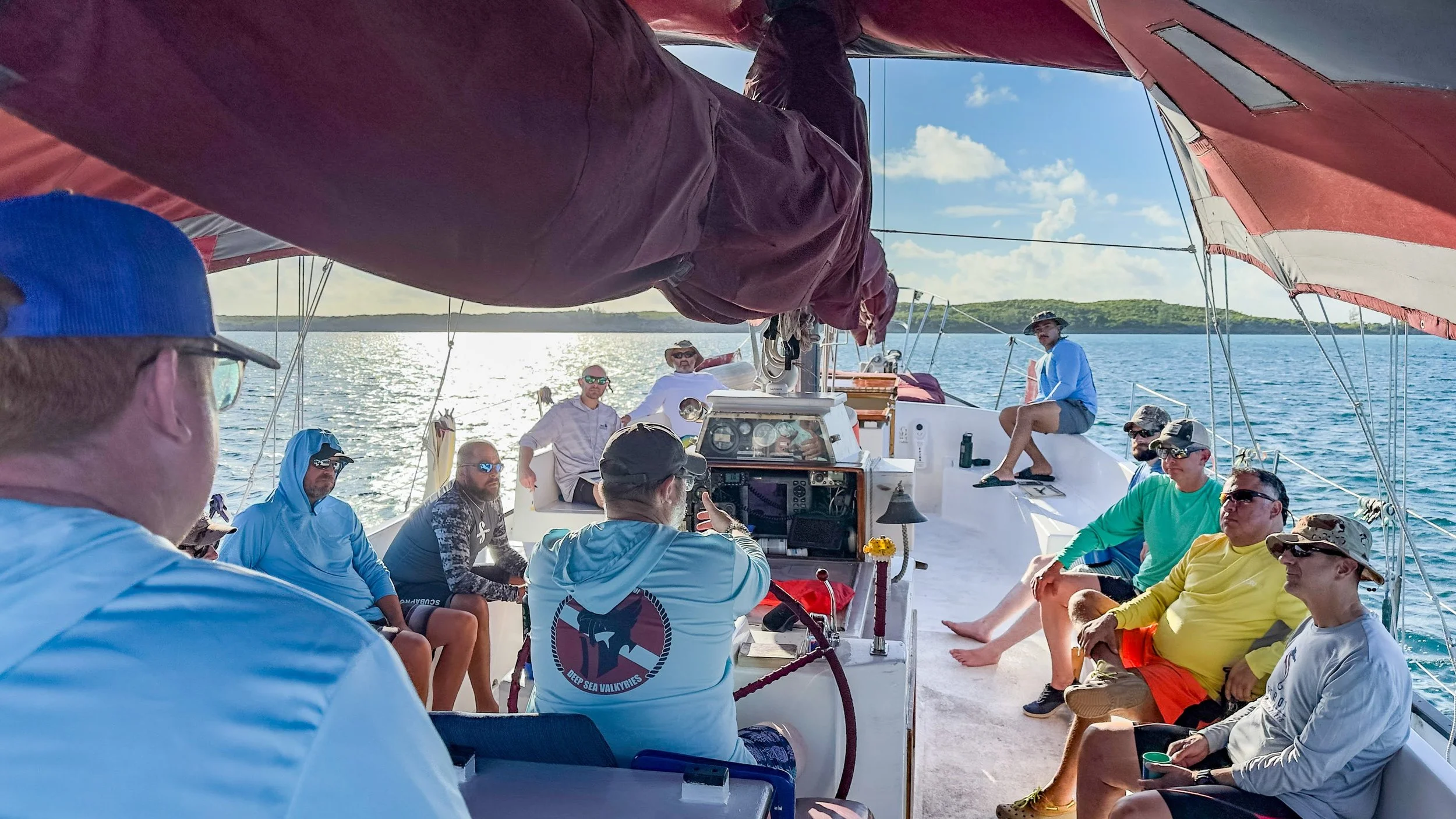 Group of people on a boat, some sitting and some standing, wearing casual attire including hats and sunglasses, with water and a distant shoreline visible in the background during sunny weather.