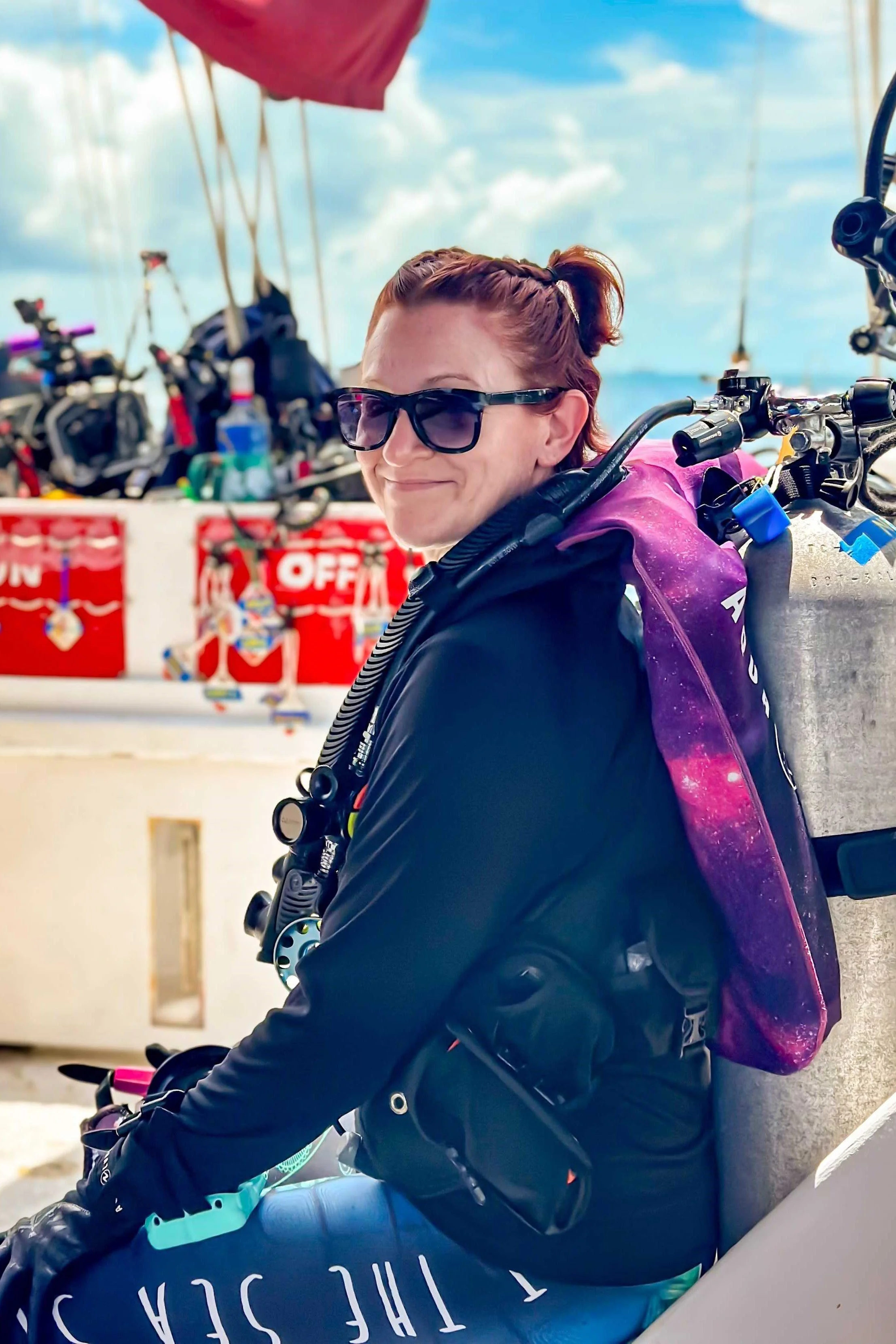 A woman with red hair and sunglasses sitting on a boat with scuba gear, smiling, with a clear sky in the background.
