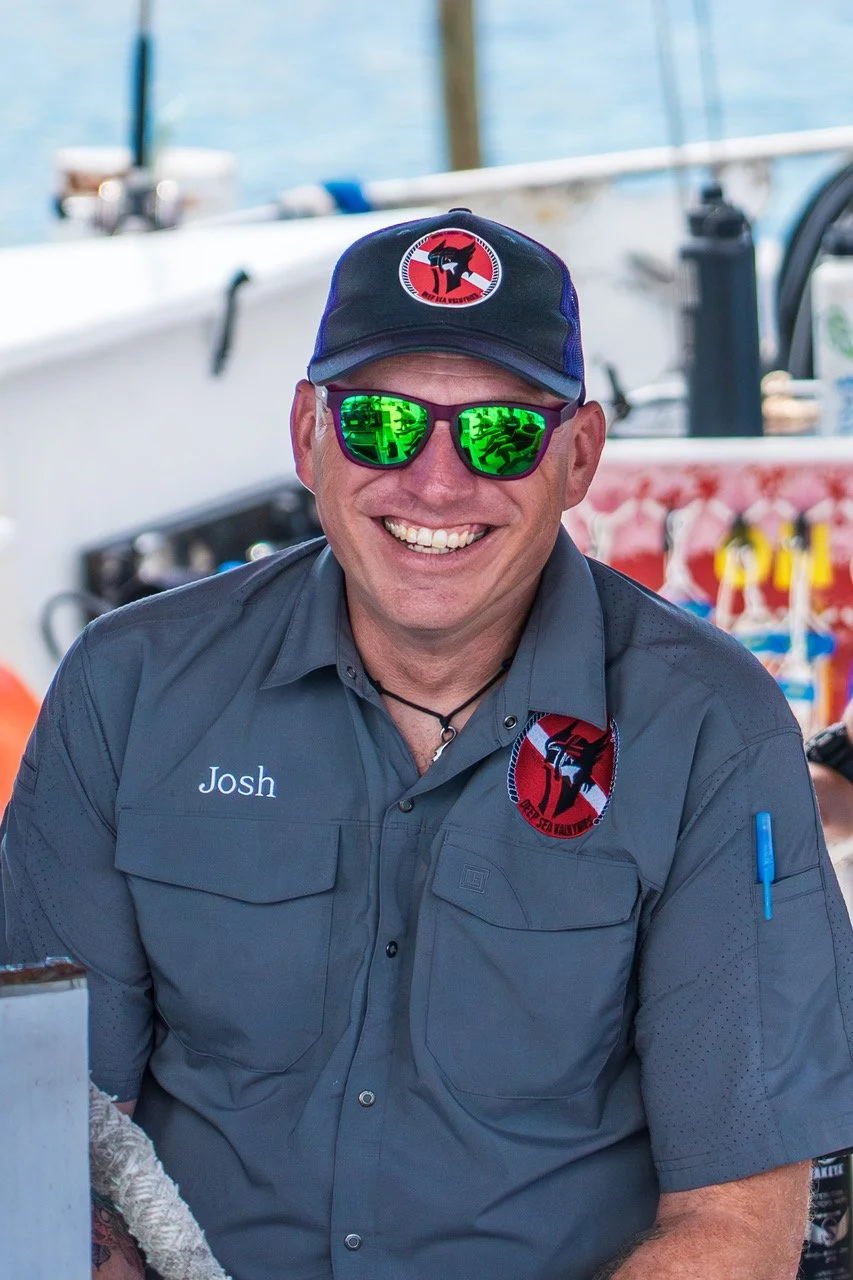 A smiling man wearing sunglasses, a navy cap with a logo, and a gray shirt with the name tag 'Josh' is at a marina near boats and water.
