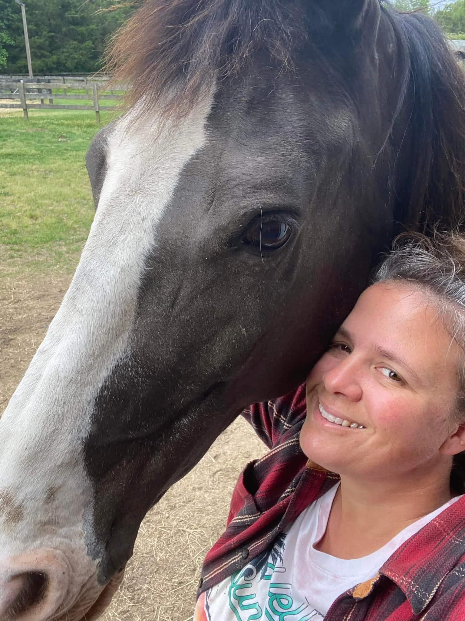 A woman smiling and leaning in close to a large black and white horse in a field with a wooden fence in the background.