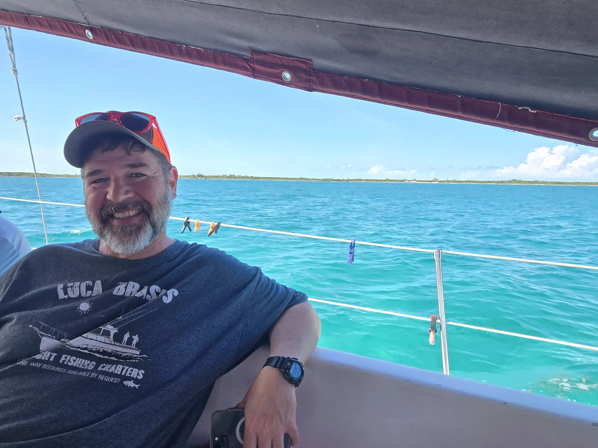 A smiling man with a beard and mustache wearing a black T-shirt, red sunglasses on his cap, and a wristwatch, sitting on a boat with clear turquoise water and a blue sky in the background.