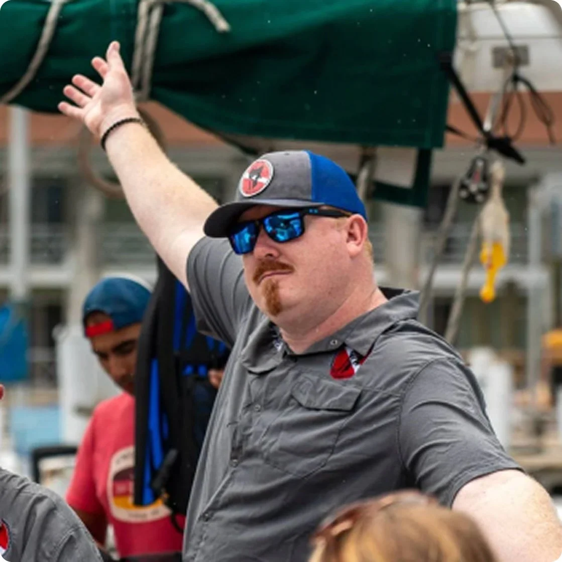 A man with a beard and mustache wearing sunglasses, a baseball cap, and a gray shirt standing outdoors with his right arm raised and pointing to the left.