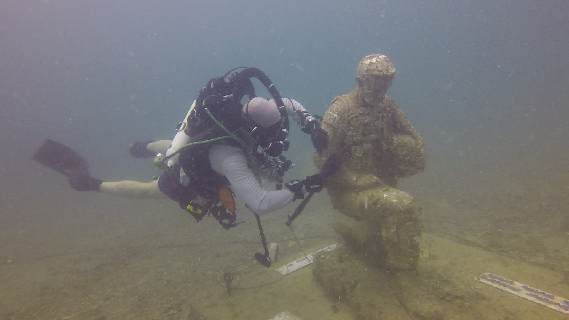 A scuba diver examining and touching a large submerged statue of a seated figure holding a book underwater.