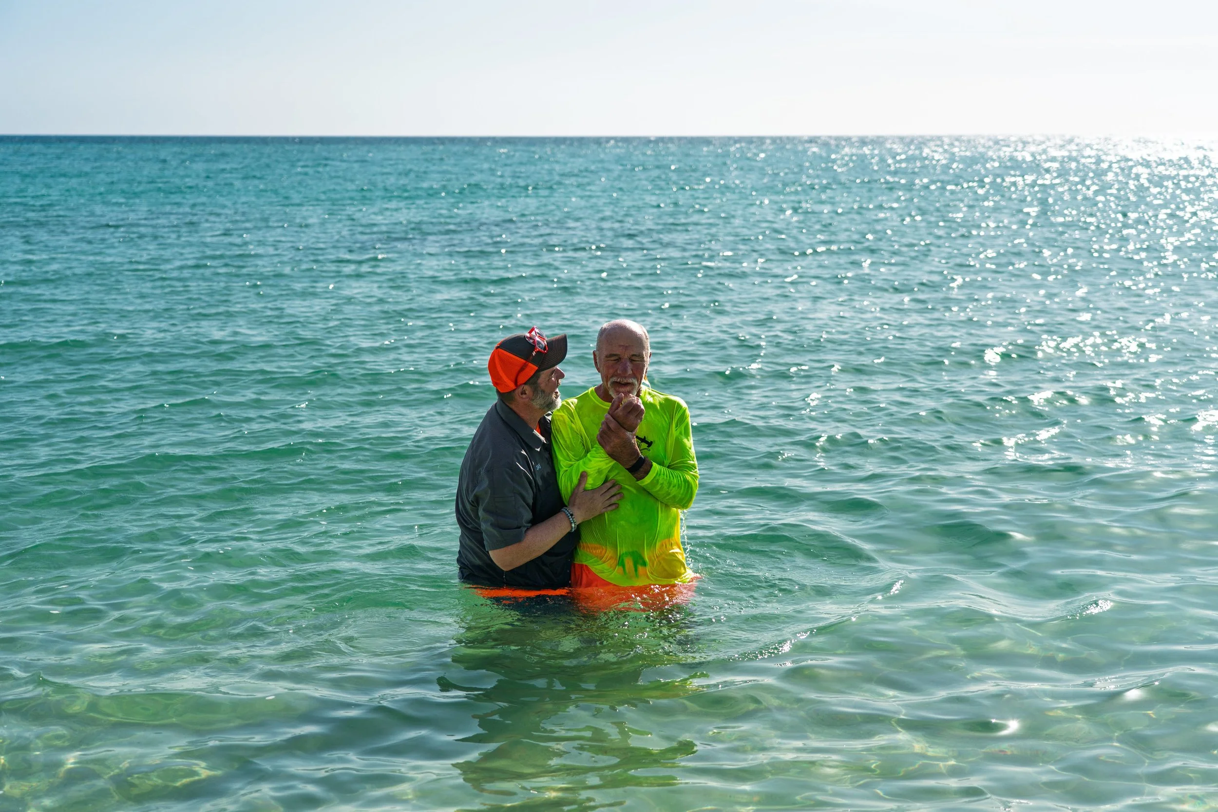 Two men stand in shallow clear ocean water, one as a priest and the other as a groom, smiling and holding hands during a wedding ceremony in the sea.