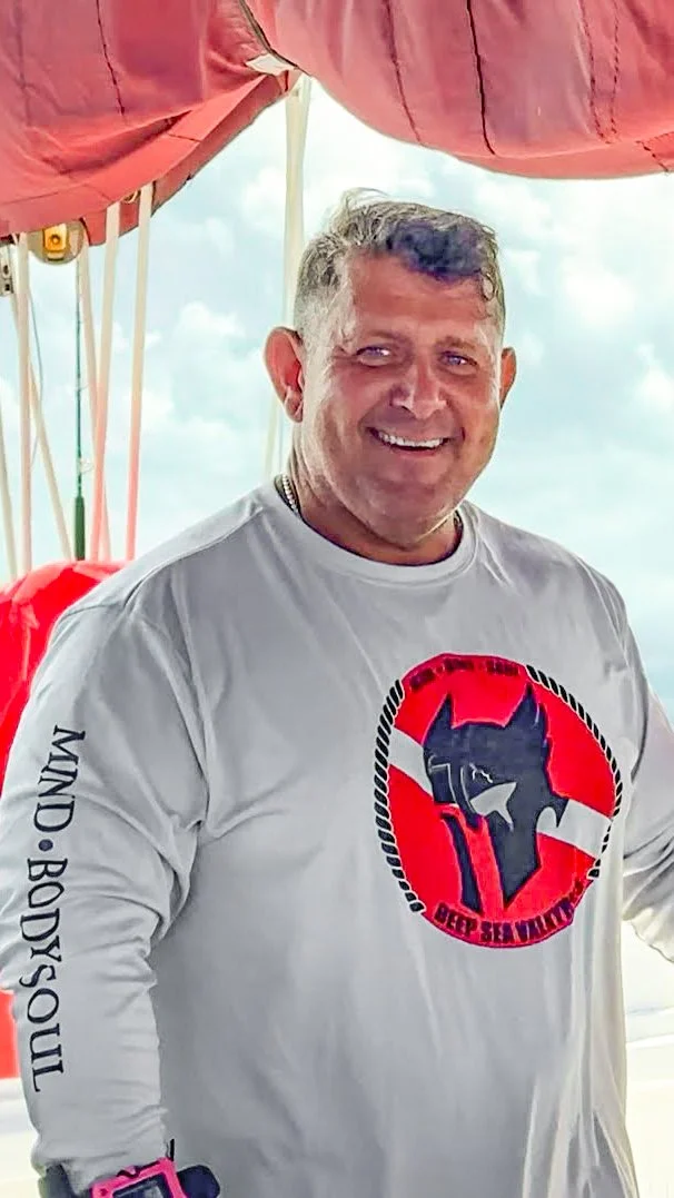 A smiling man with short gray hair outdoors under a pink canopy, wearing a light gray long-sleeve shirt with a logo featuring a blue wolf head and the text "BEP SEAWOLF" on the front, and a black and white logo with the head of a knight's helmet on the chest. The shirt's left sleeve has the words "KIND • BODY• SOUL." He's also wearing a black wristband and a pink watch.