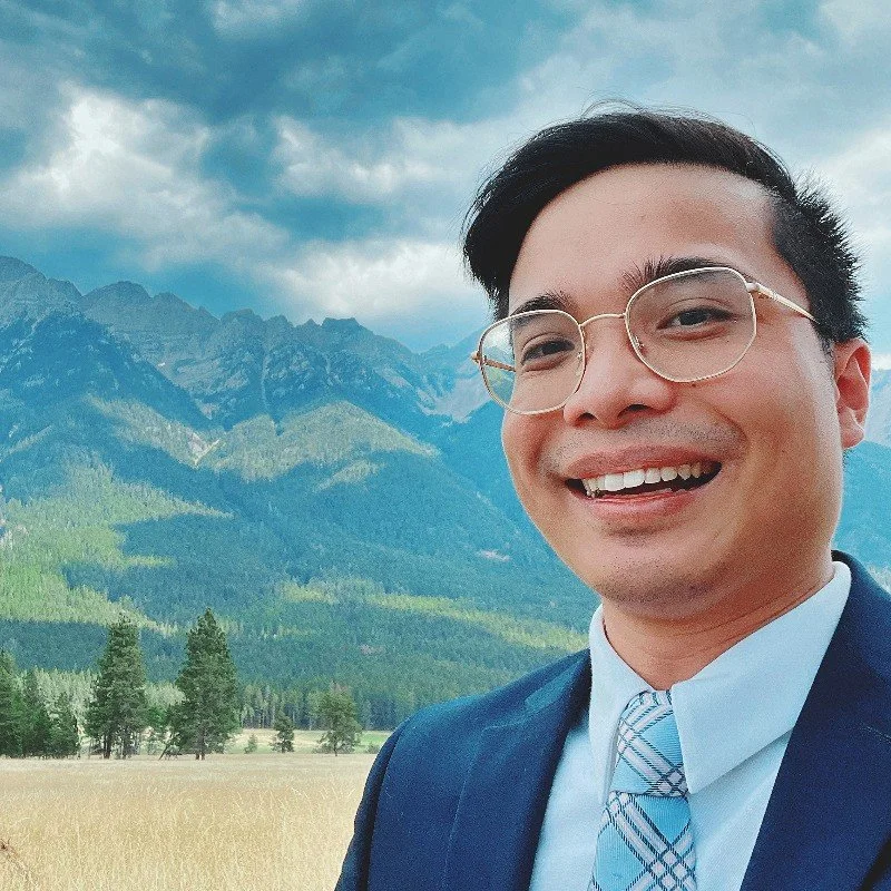 A smiling man in glasses and a suit stands outdoors in front of mountains and a cloudy sky.