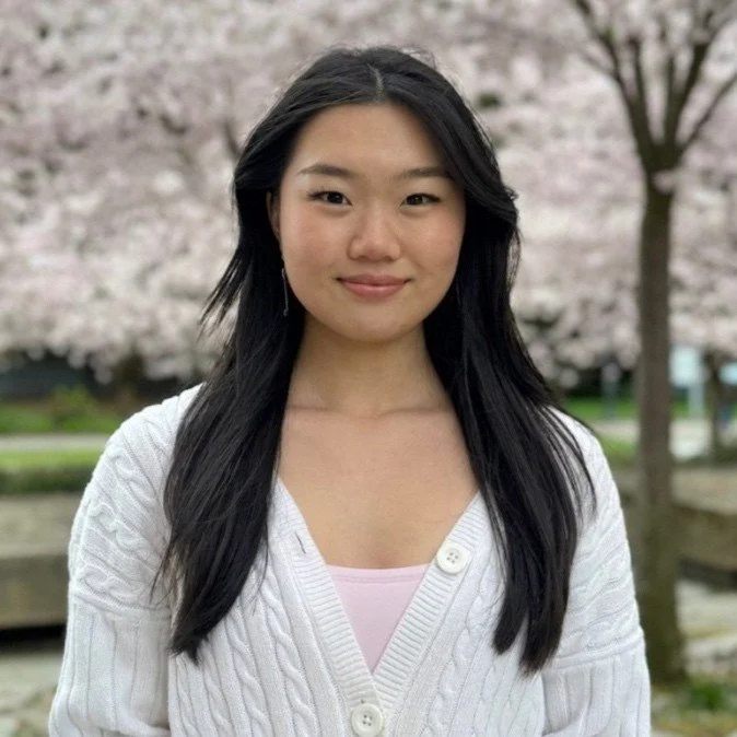 A young woman with long black hair, smiling, standing outdoors in front of blooming pink cherry blossom trees.