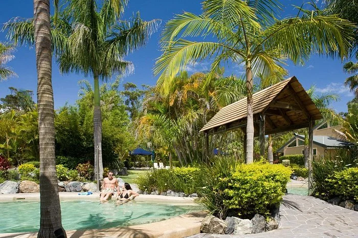Schoolies holiday. A tropical pool scene with palm trees, a small pool with people relaxing, and a shaded wooden structure in the background on a sunny day.