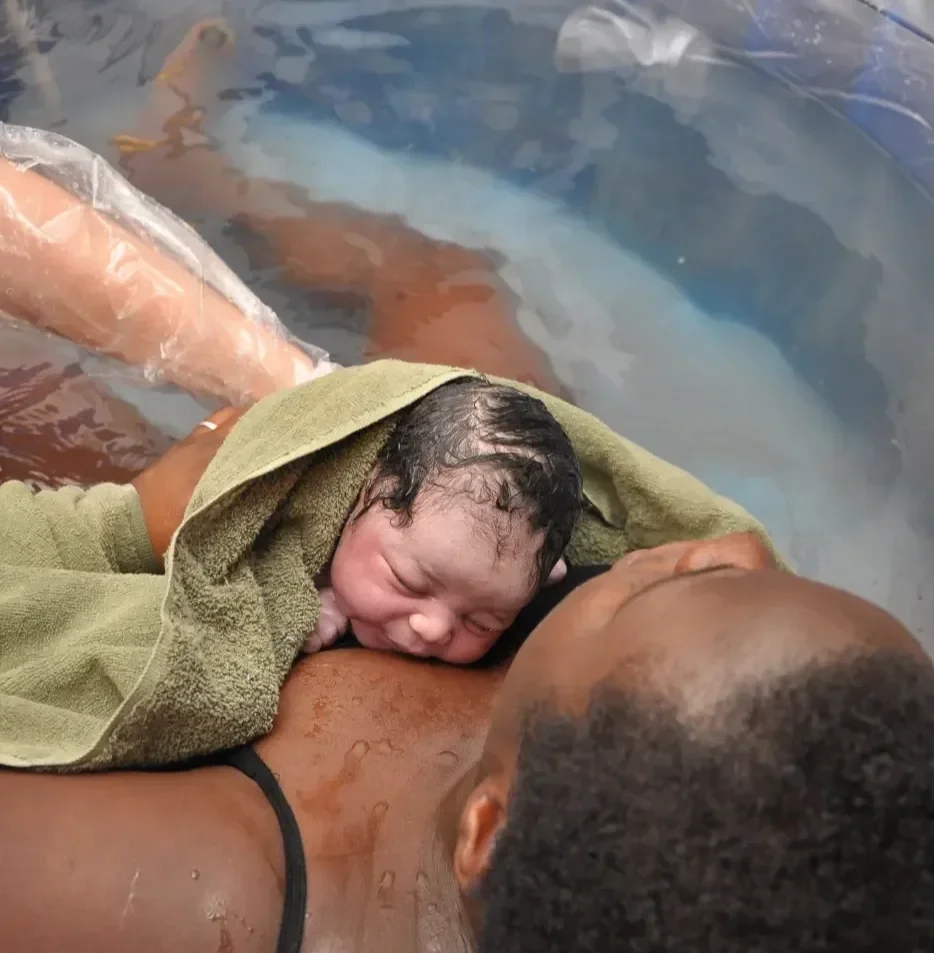 A newborn baby with dark, wet hair resting on an adult's chest in a water setting, with another person in the background underwater, partially visible.