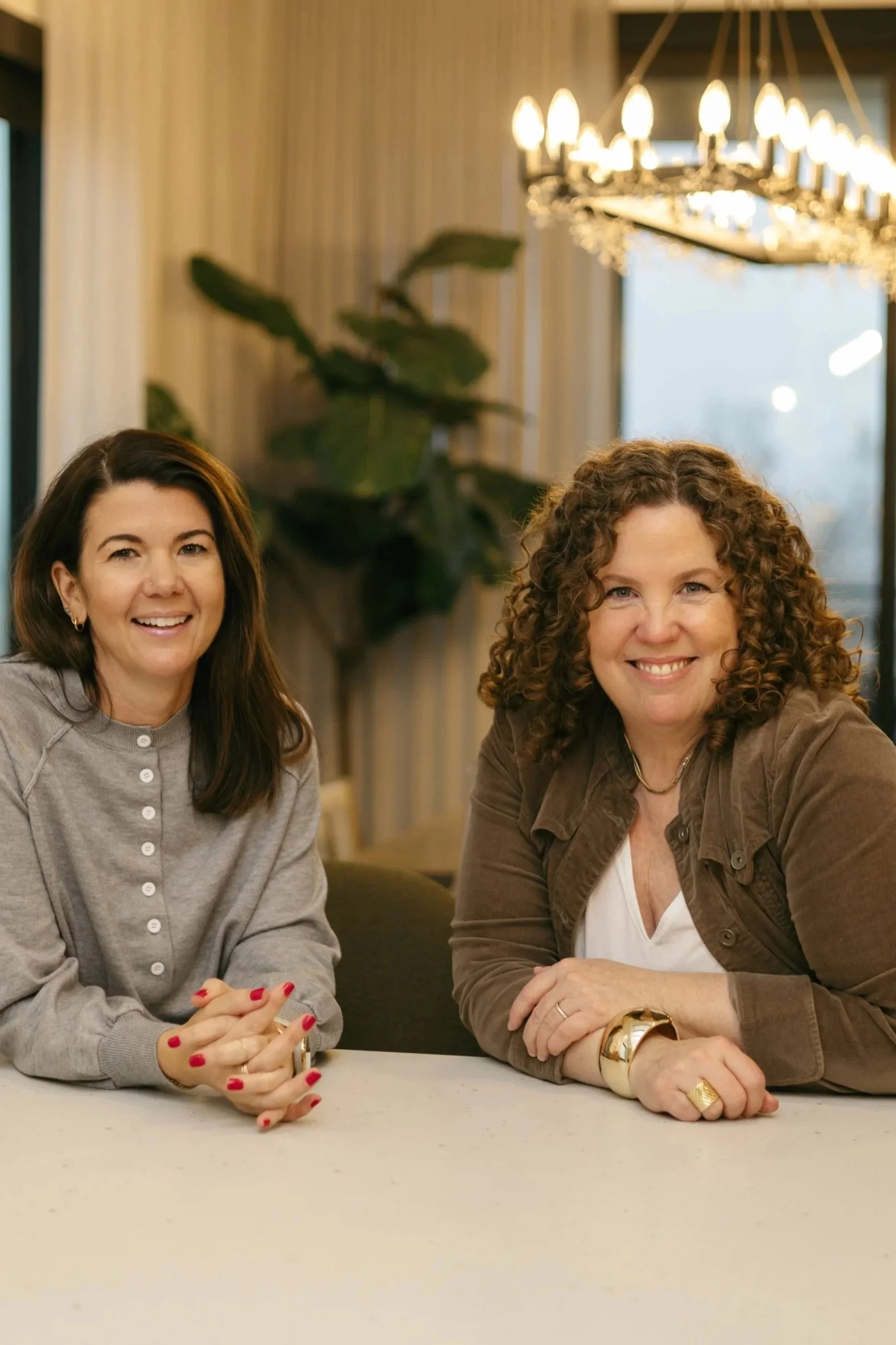 Two smiling women sitting at a table in a warmly lit room with a chandelier and large window in the background. One woman has dark brown hair, wearing a gray cardigan. The other woman has curly red hair, wearing a brown jacket and jewelry.