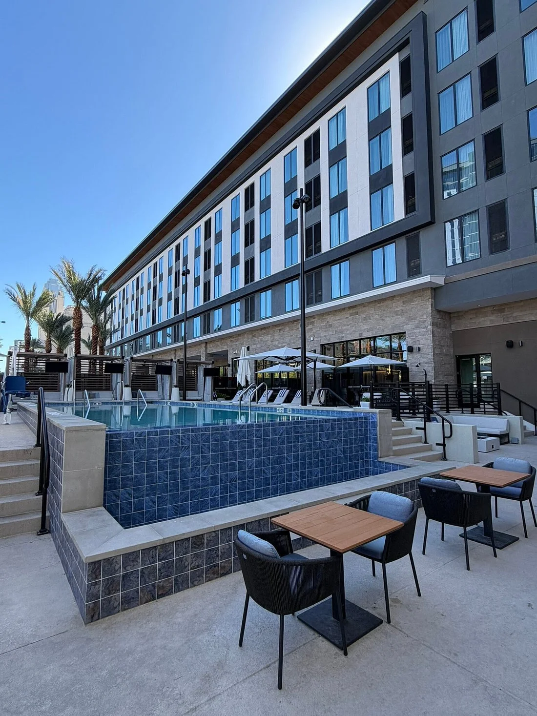 Modern multi-story building with large blue window panes and beige brick accents under a clear blue sky, with palm trees and outdoor seating area.