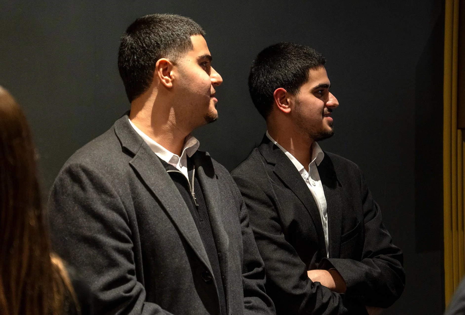 Two men in formal attire standing side by side against a dark background.
