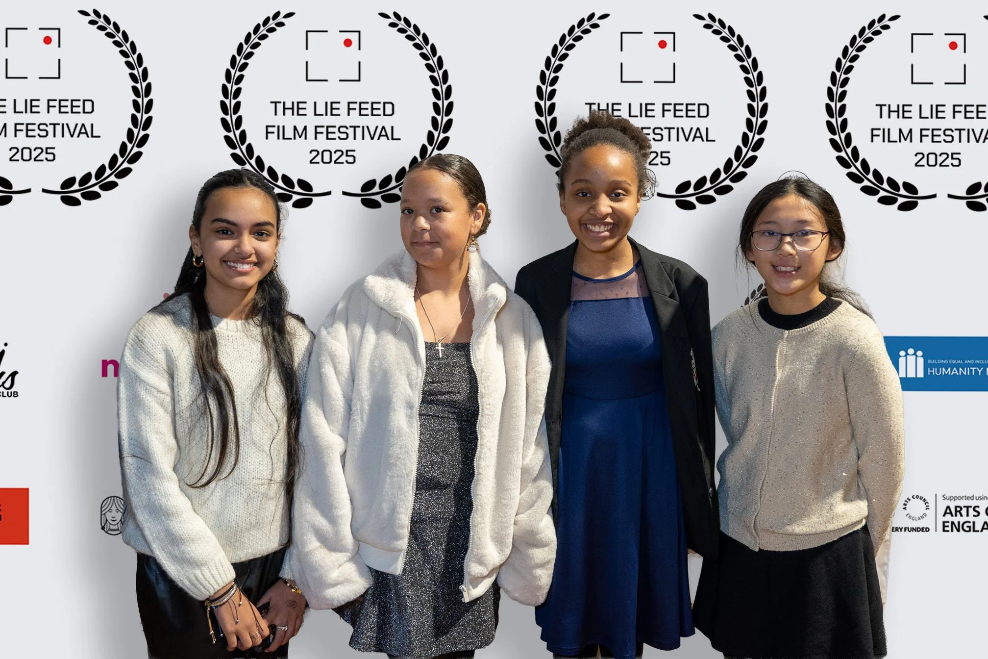 Four girls standing in front of a white backdrop with black laurel wreath logos and text that reads "The Lie Feed Film Festival 2025." They are smiling and posing for the photo.