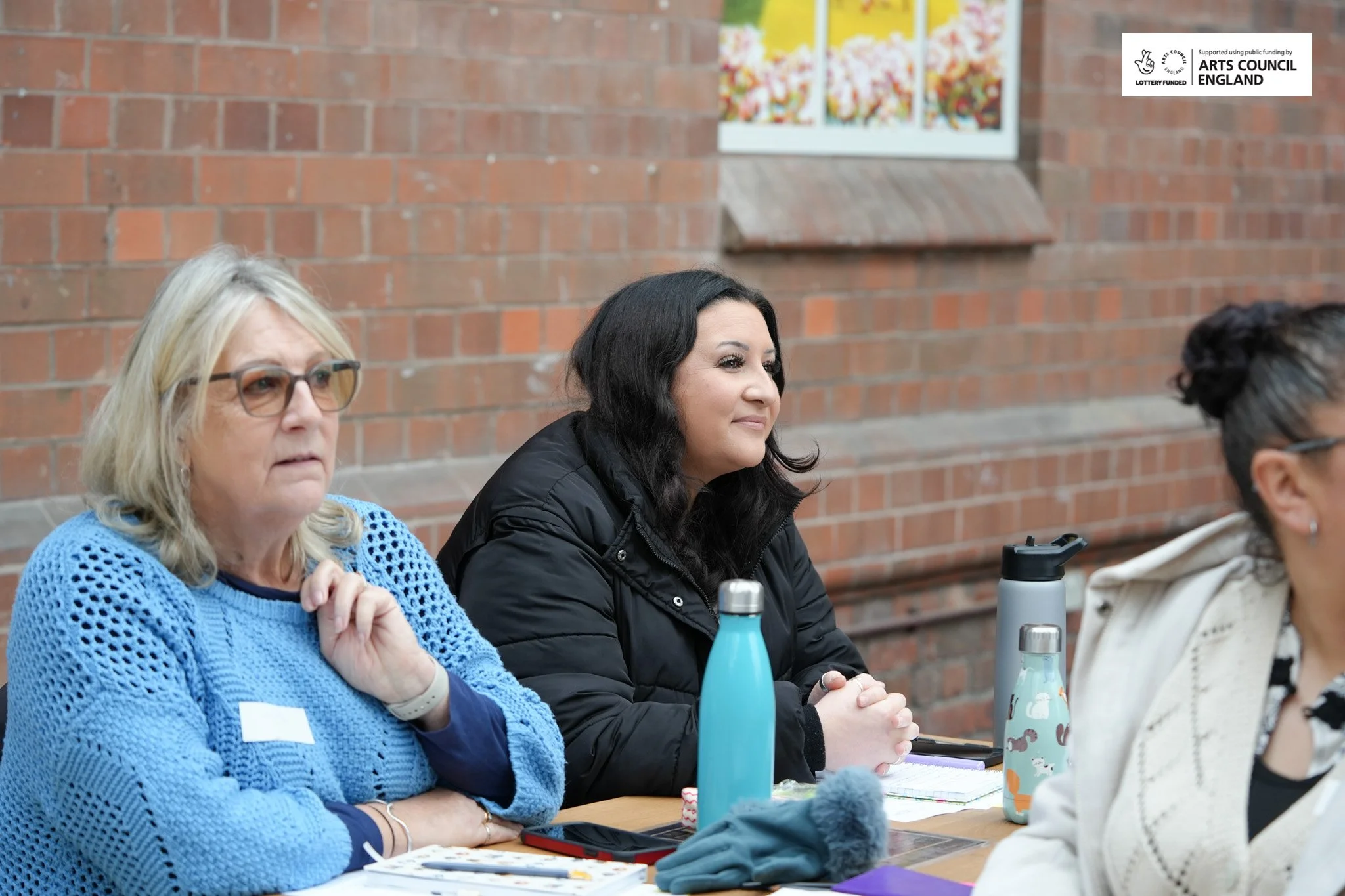 Three women sitting at a table during a meeting or workshop, with water bottles and notebooks in front of them, against a brick wall background.