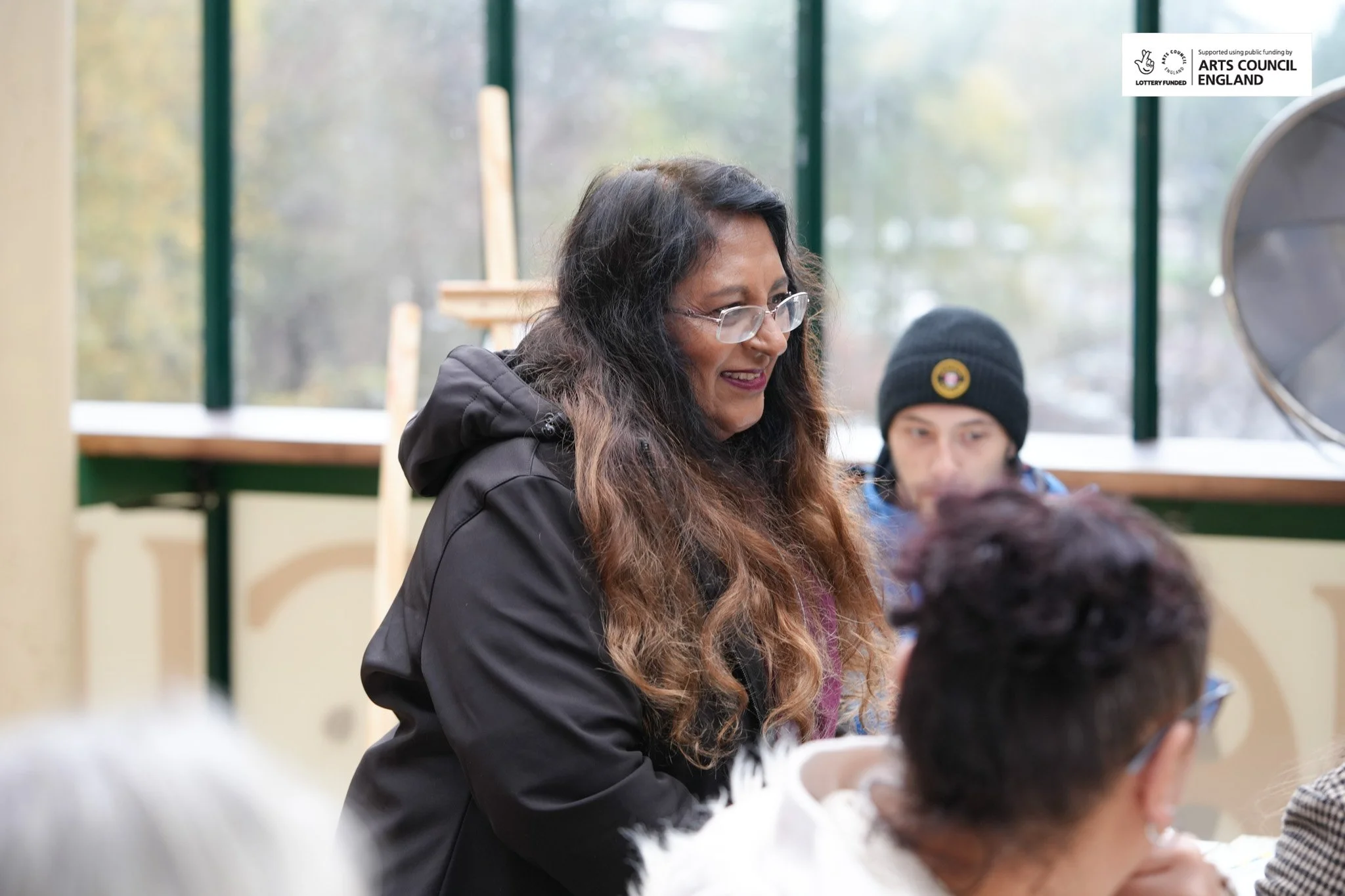 A woman with long wavy hair, glasses, and a black jacket smiling and talking with two other women in an indoor setting with large windows and natural light.