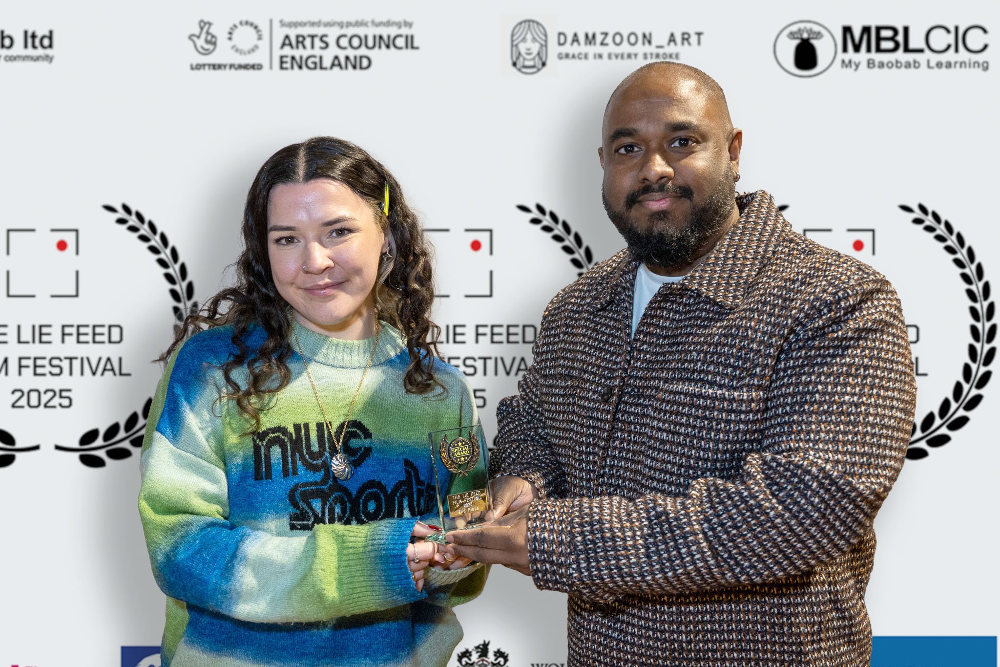 A woman and a man stand side by side, holding an award together at the Lie Feed Film Festival 2025, in front of a backdrop with various logos and text.