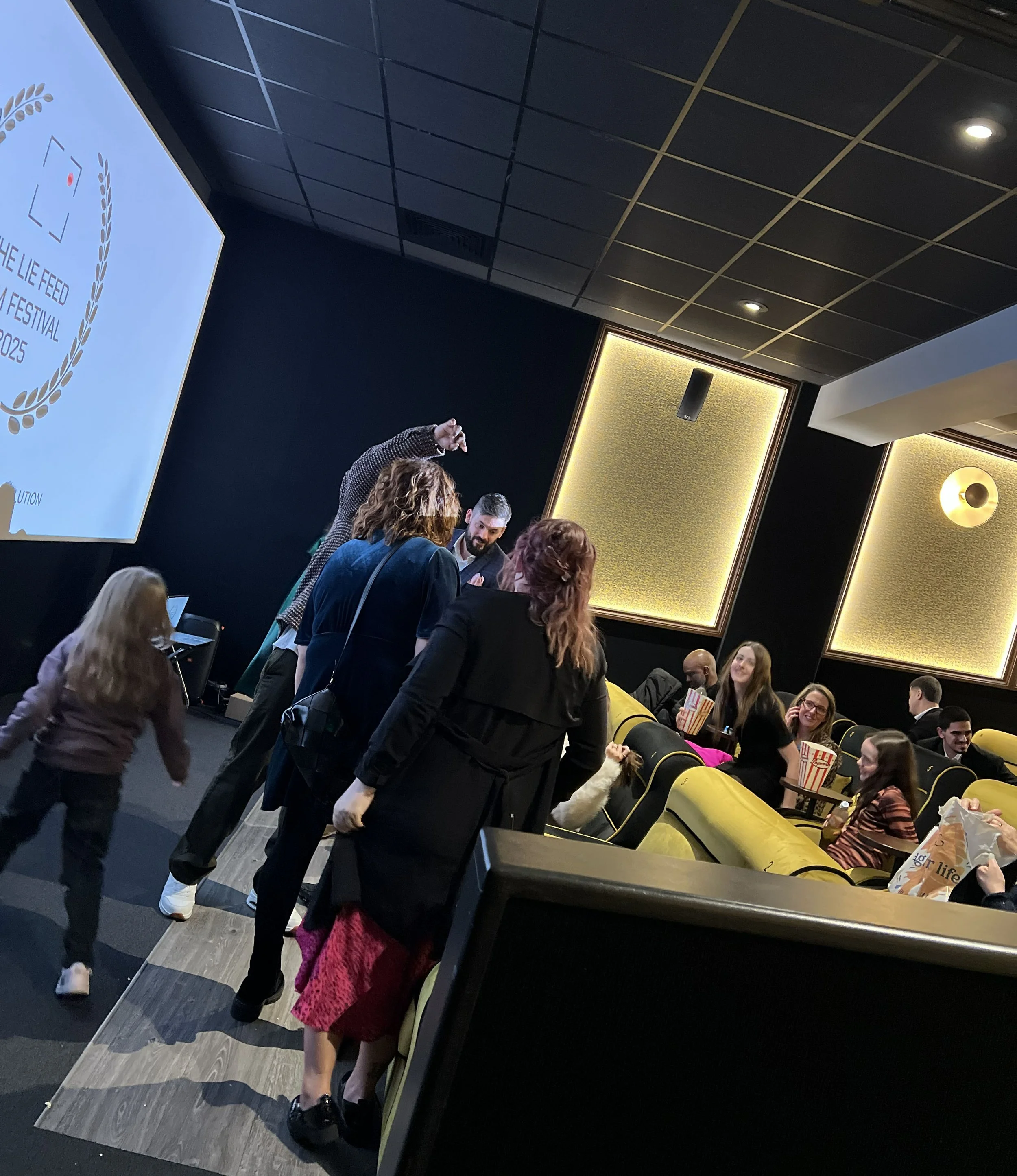 People gathering in a dark movie theater, some seated on yellow seats and others standing, with a large screen displaying text on the left side.