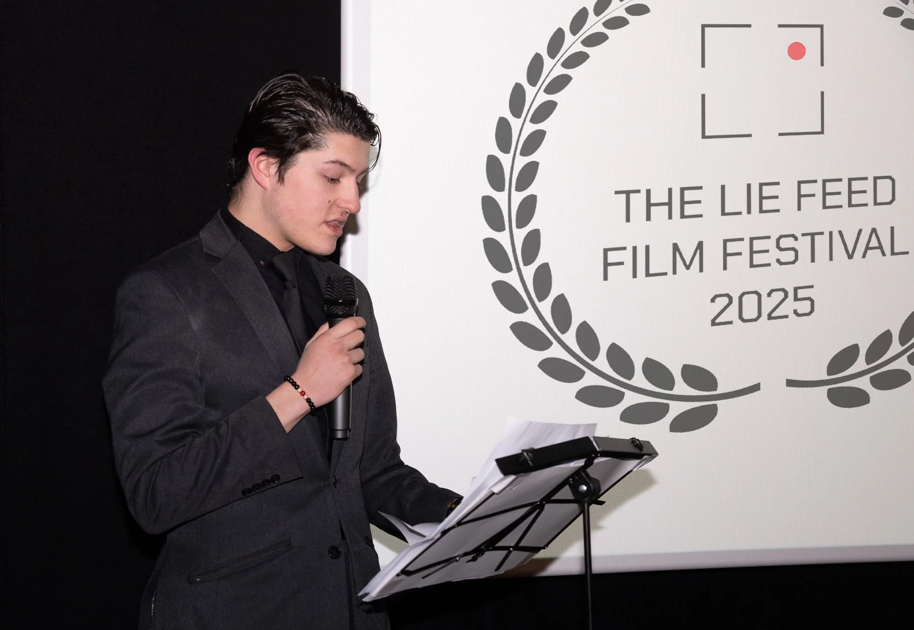 A young person with short dark hair, wearing a dark suit and holding a microphone, stands at a lectern with papers, speaking at the Lie Feed Film Festival 2025 event.