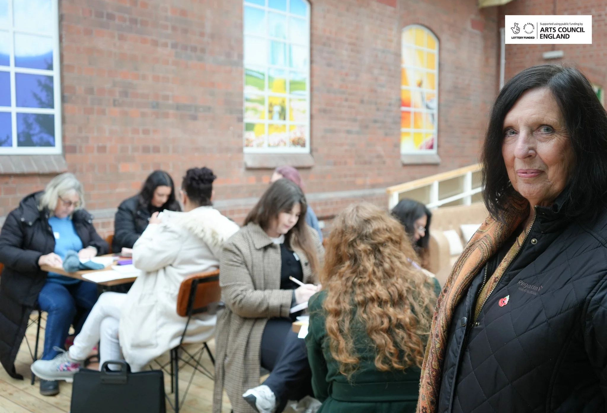 A group of diverse women sitting at tables in a room with brick walls and stained glass windows, engaged in writing or reading. A woman with dark hair stands in the foreground, smiling at the camera.