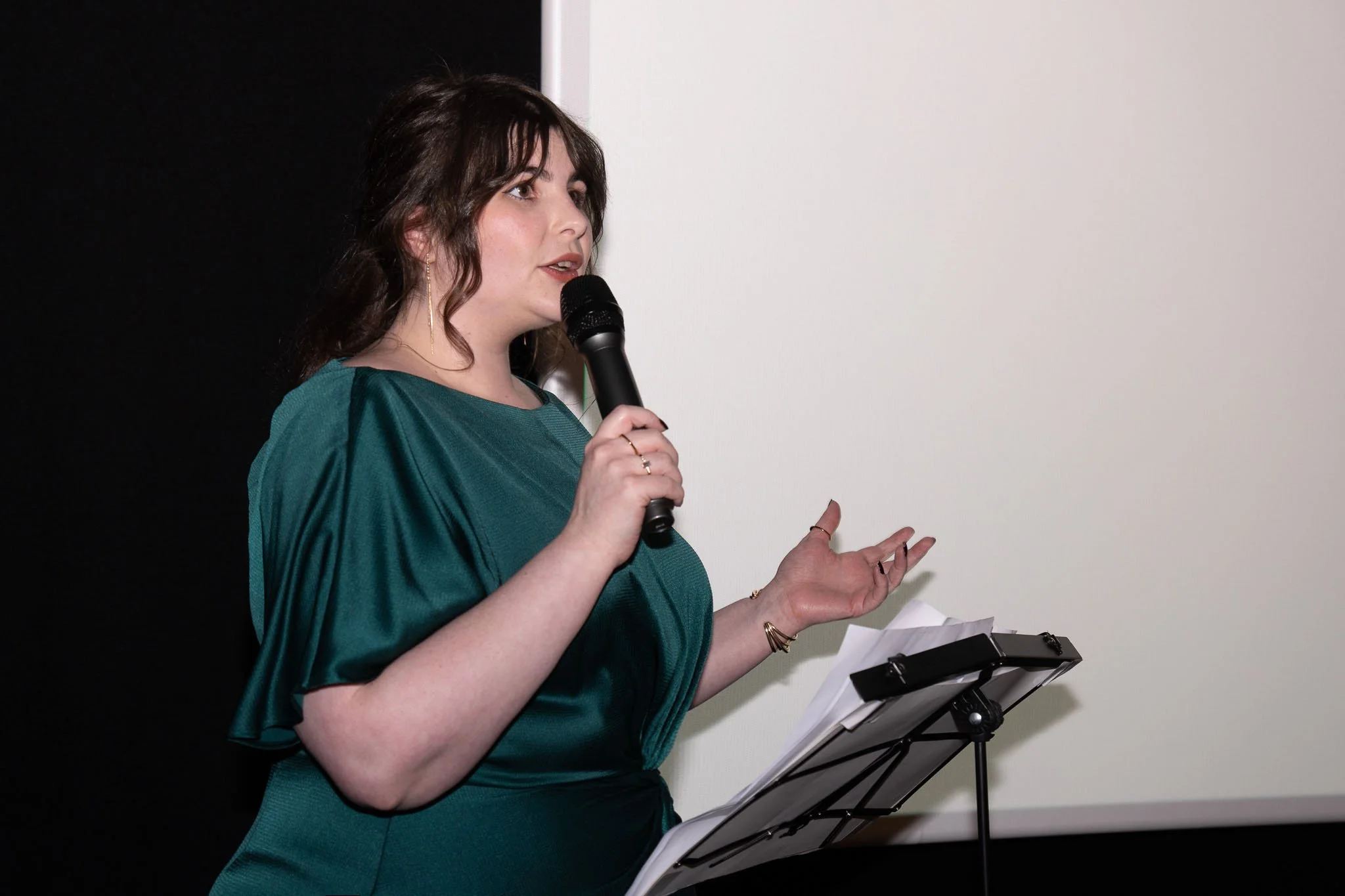 A woman with dark brown hair, wearing a teal dress, speaking into a microphone and gesturing with her hand while standing at a music stand with papers.