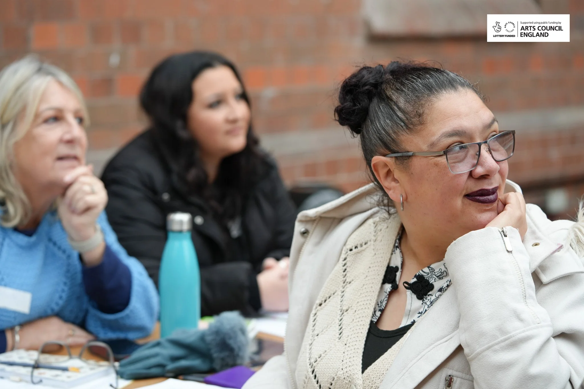 Three women sitting at a table during a discussion or meeting indoors, with a brick wall in the background. The woman closest to the camera has glasses, dark hair in a bun, and is wearing a cream-colored jacket. The middle woman has dark hair and is 