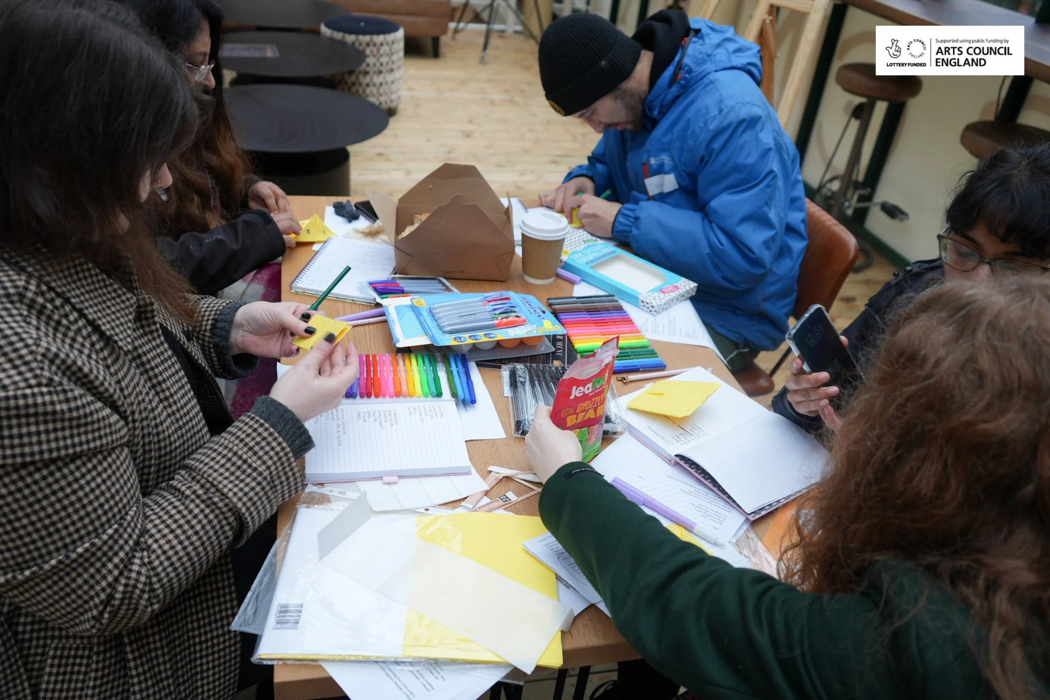 Group of people sitting around a table filled with colorful pens, notebooks, and stationery, engaged in an arts and craft activity, with a sticker of the Arts Council England logo in the top right corner.