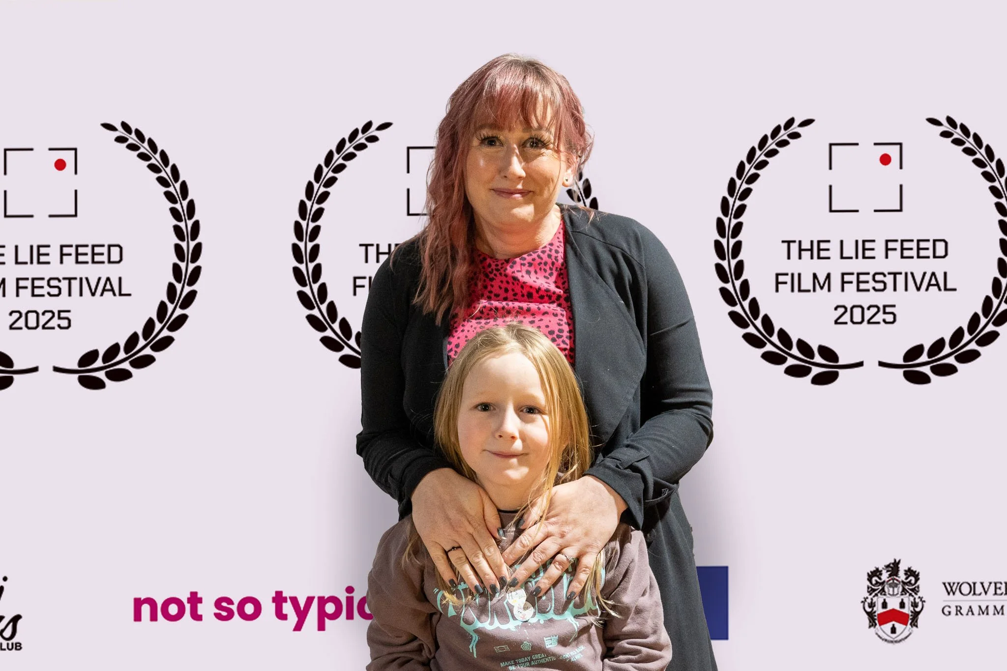 A woman and a young girl standing in front of a backdrop with film festival logos and text. The woman has shoulder-length pinkish hair, and the girl has long blond hair. The woman is wearing a black jacket over a pink shirt with black spots, and the 