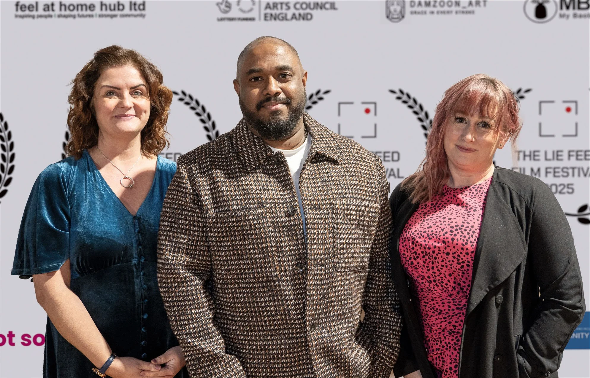 Three people standing together at a film festival event backdrop. The woman on the left has curly red hair, wearing a shiny blue dress. The man in the middle has a beard, short hair, wearing a checkered jacket. The woman on the right has wavy pink ha