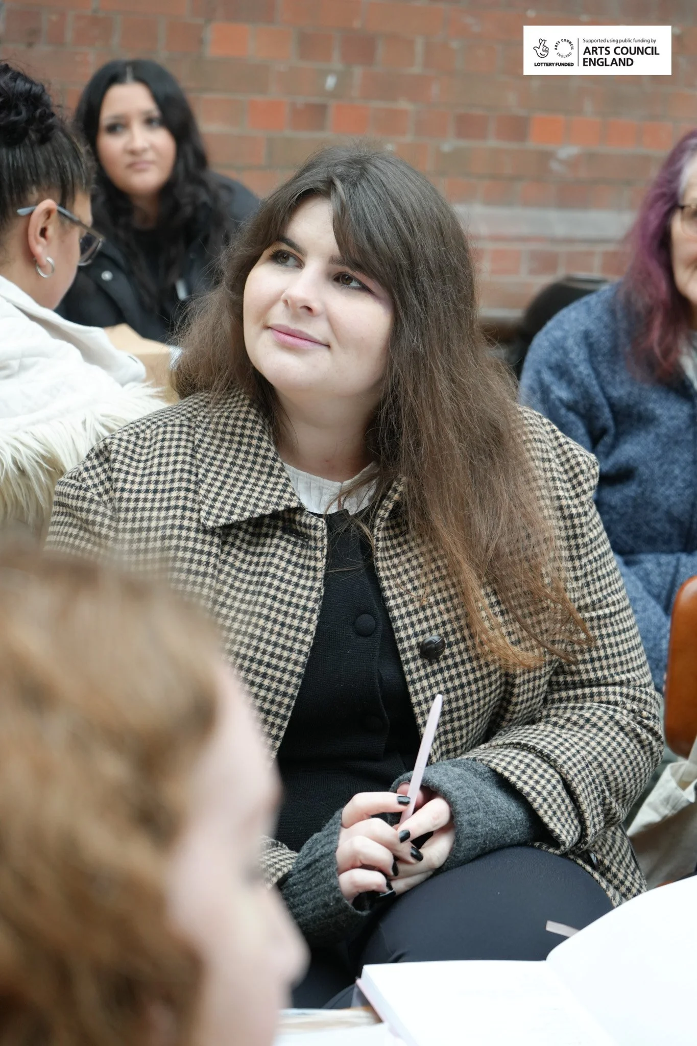 A woman with long brown hair wearing a checkered coat, sitting at a table, holding a pen, and listening attentively at an indoor event with a brick wall background.