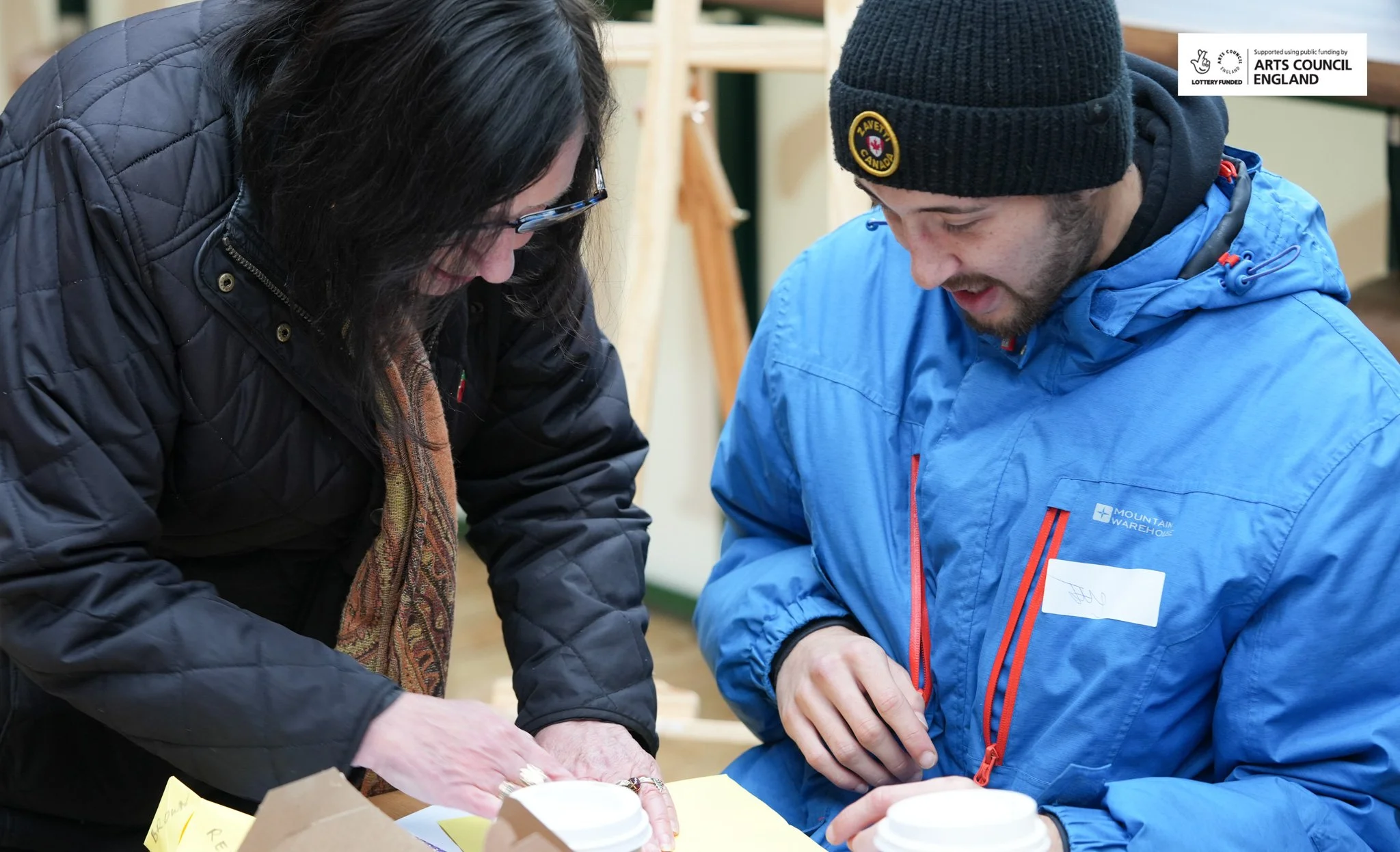 Two people, a woman and a man, are looking at something on a table and smiling, indoors.