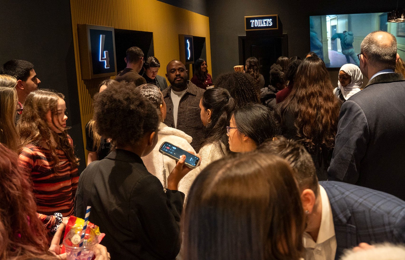 Group of diverse people gathered in a busy indoor space, some looking at a large screen television showing a dog, with signs indicating the number 4 and 3 and a sign for toilets visible on the wall.