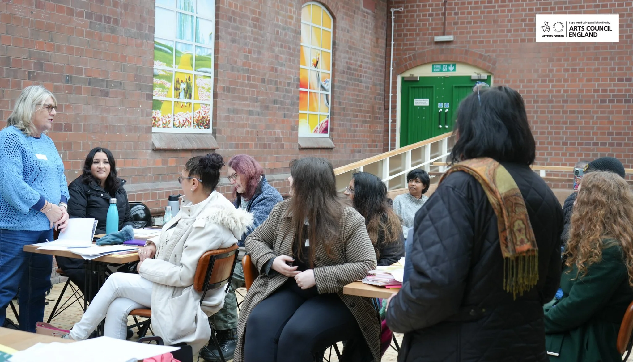A group of diverse women engaged in a discussion or workshop in a room with brick walls, large windows with colorful floral and sunset murals, and a green door in the background.
