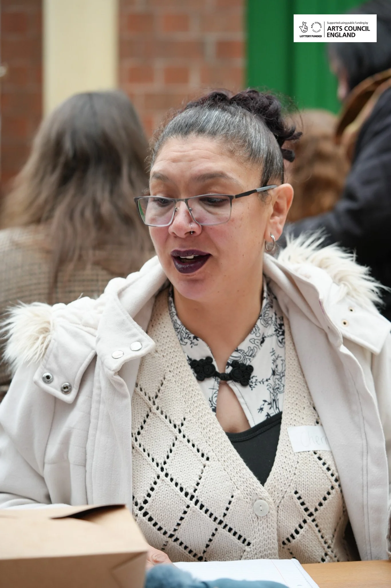 A woman with glasses and dark lipstick at a table, wearing a light-colored sweater vest and a white coat with a fur collar, at a social or community event, with other people in the background.