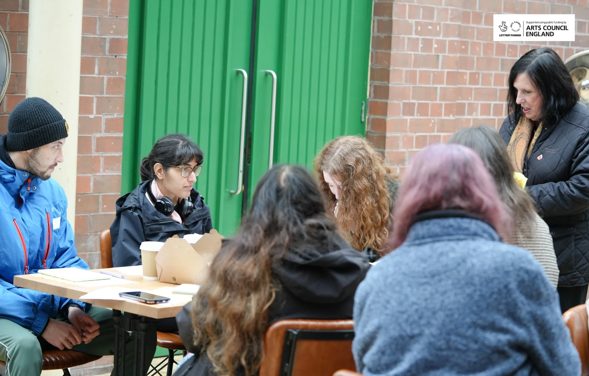 A group of people sitting around a table in an indoor setting, with a brick wall and green door in the background. A woman standing appears to be addressing or speaking to the group.