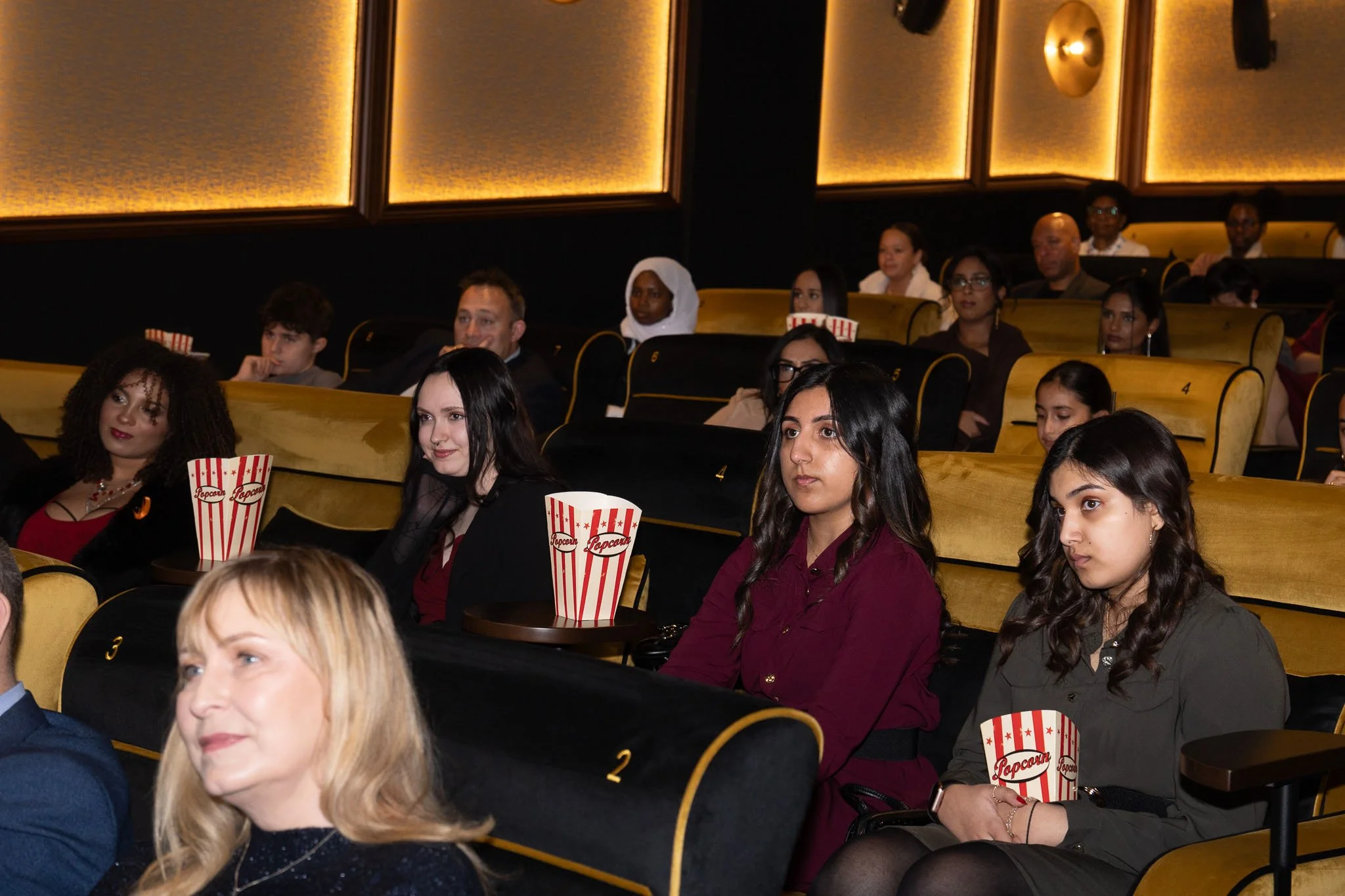 People seated in a theater with yellow and black seats, watching a movie or a presentation, with some holding red and white striped popcorn containers.