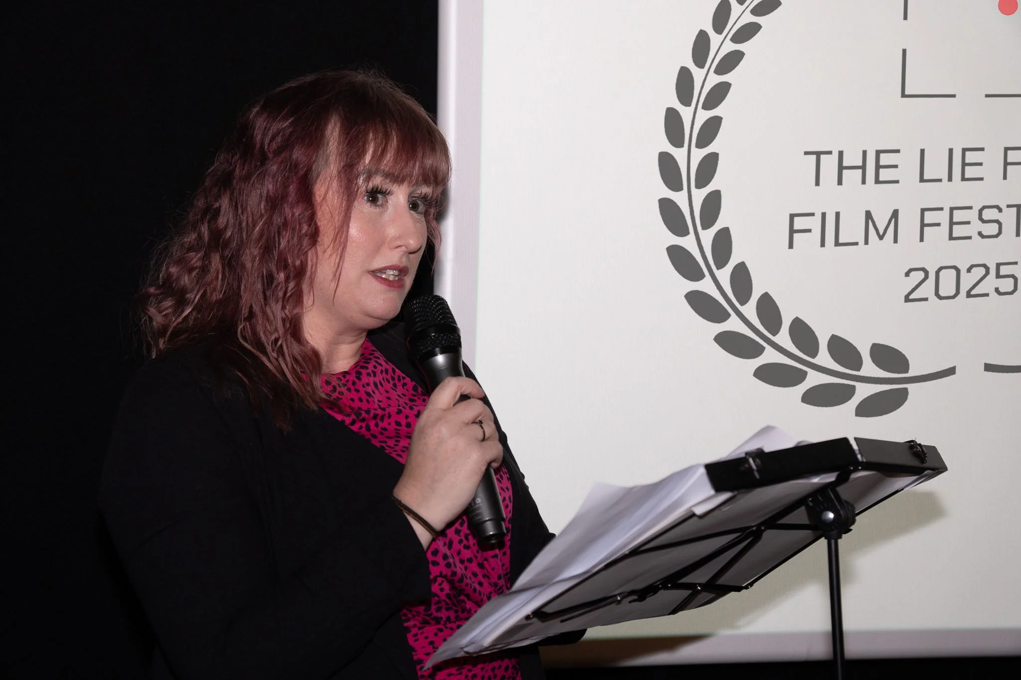 A woman with wavy, reddish hair is speaking into a microphone while reading from papers on a black music stand. She is wearing a pink and black animal print top and a black jacket. Behind her, there is a white screen with text about the 2025 film fes