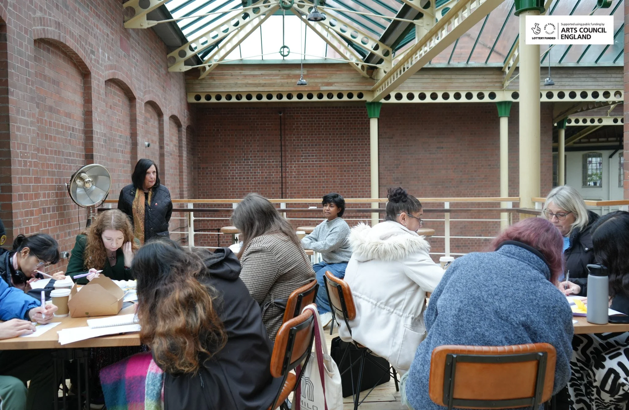 A group of women seated around a table inside a brick-walled building with a glass ceiling, engaged in work or discussion, with one woman standing and observing.