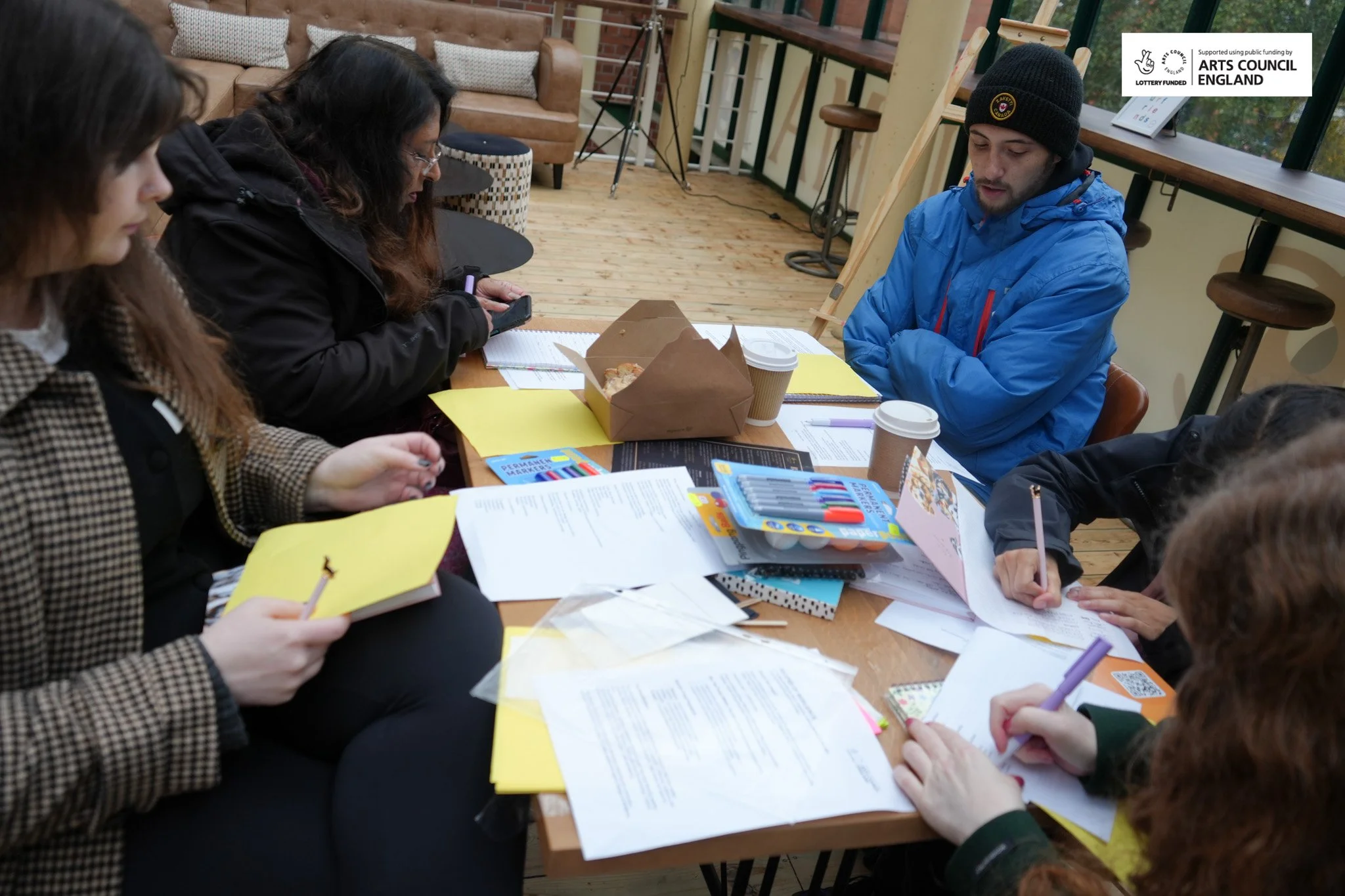 Group of people sitting around a table working on documents and notebooks with coffee cups, in a cozy cafe or meeting space with wooden floors and a sofa in the background.