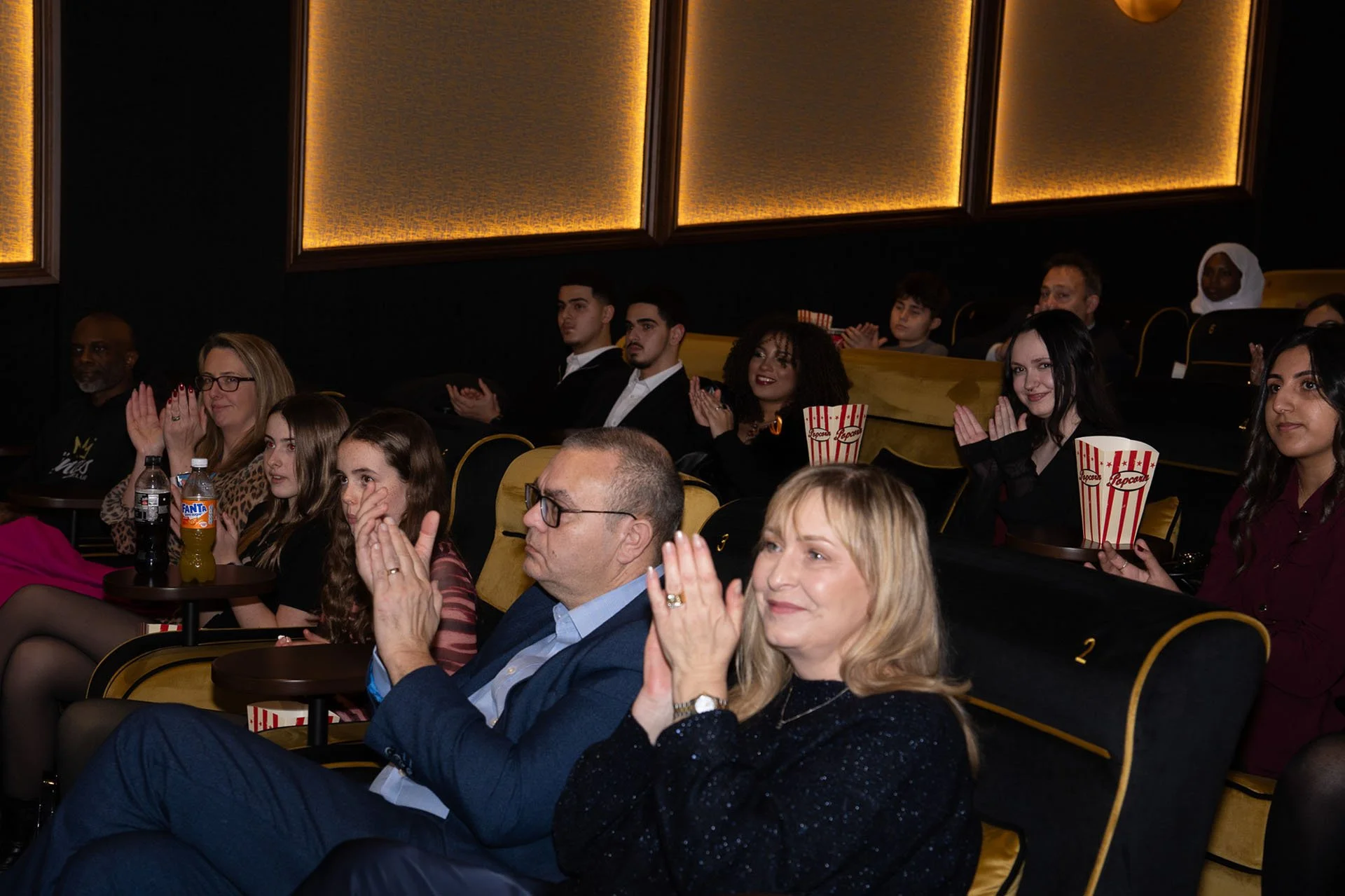 Audience seated in theater watching a performance, with some clapping and holding popcorn, in a dimly lit room with gold-colored walls.