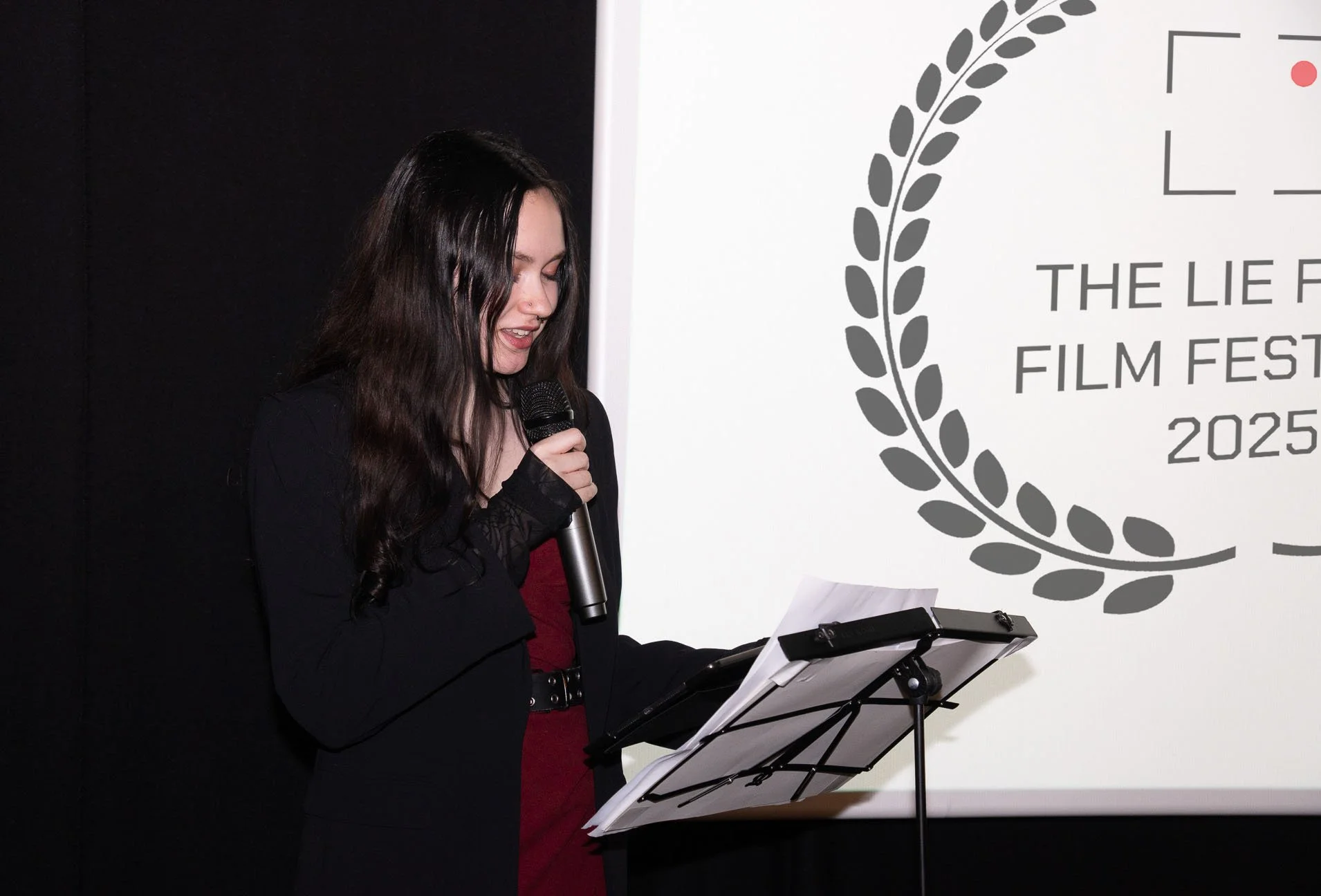 A woman with long dark hair speaking into a microphone while reading from a paper on a stand at the 2025 LIE Film Festival.