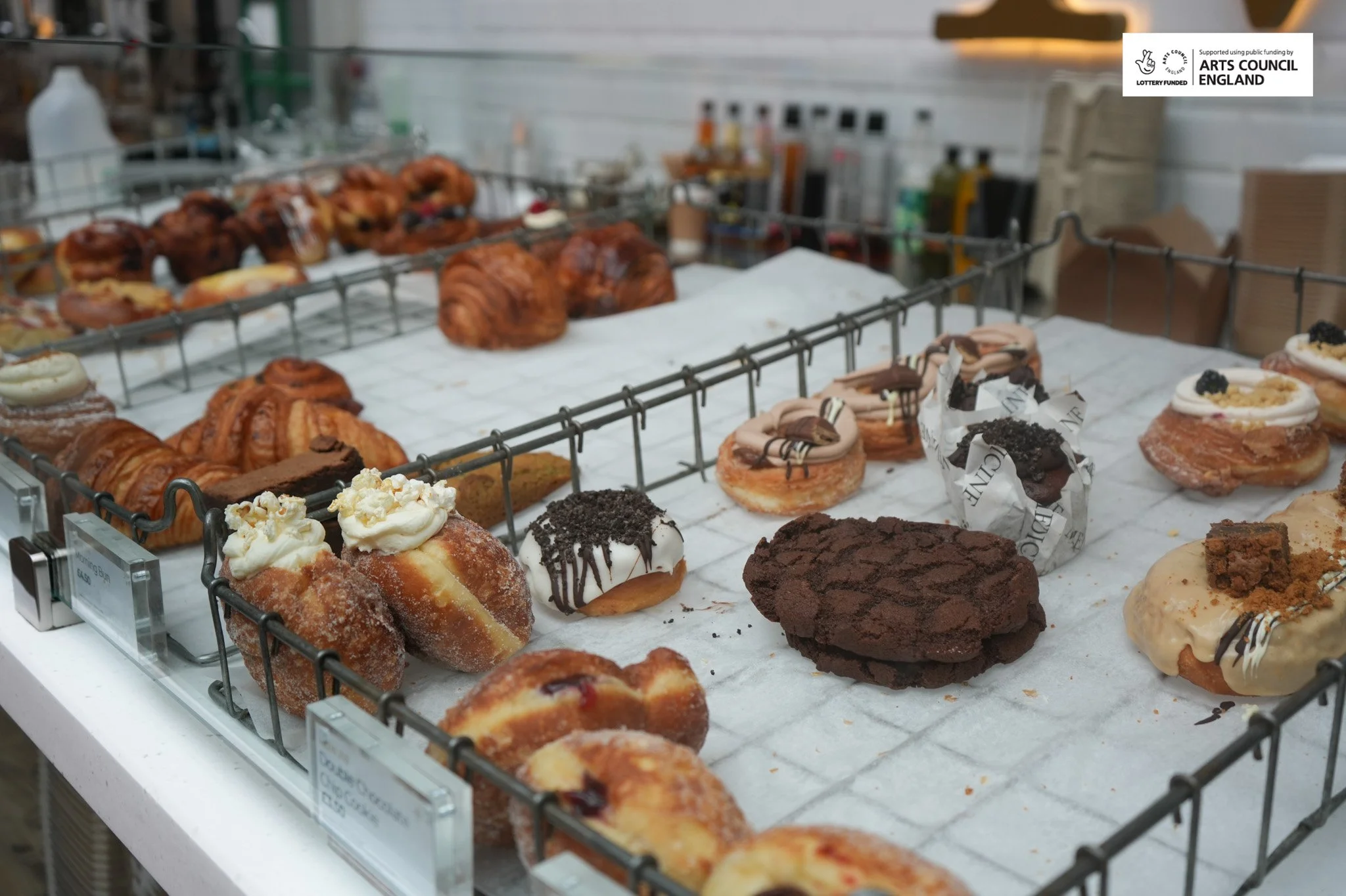 Display of various baked pastries including donuts, croissants, muffins, and cookies on a wire rack in a bakery.