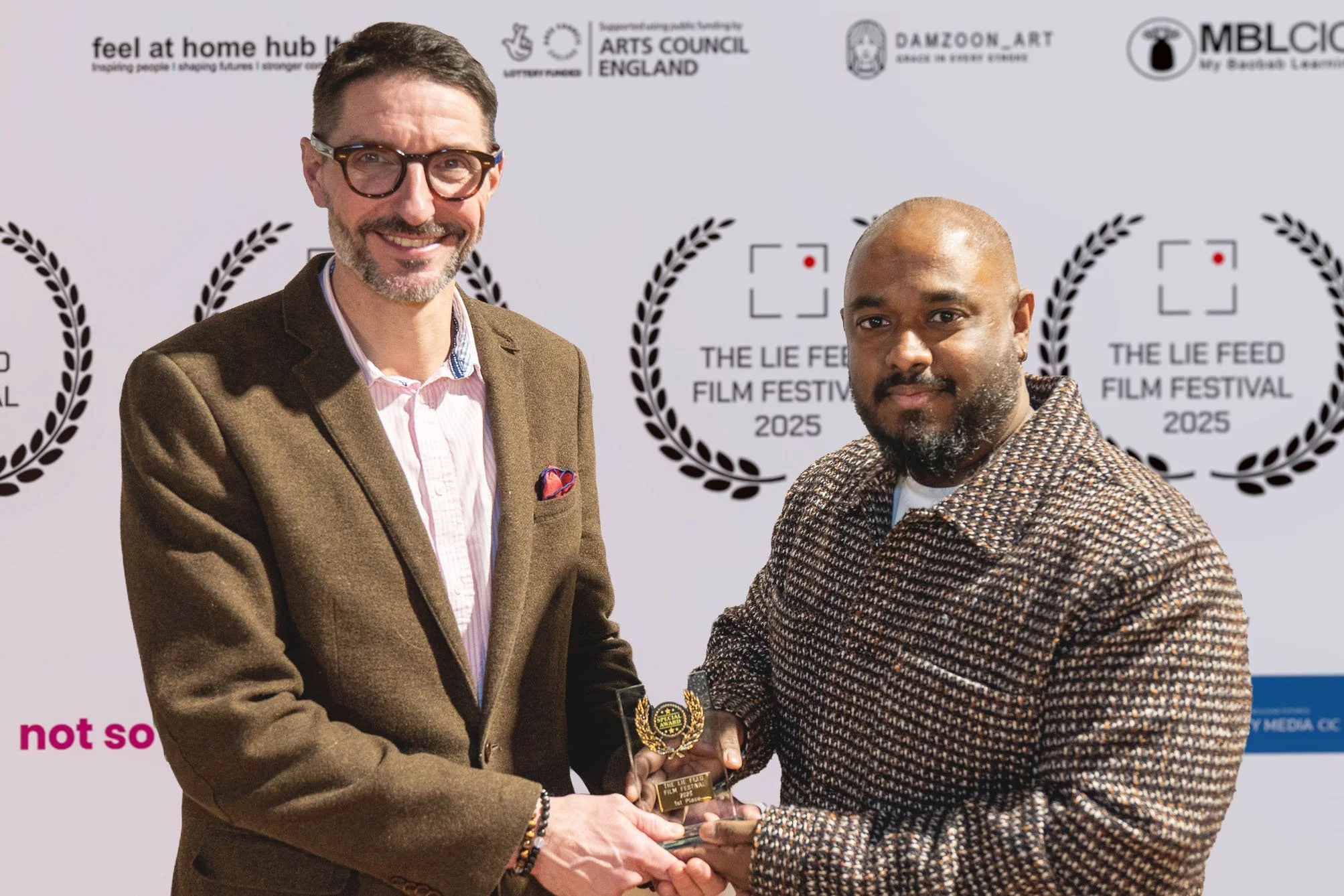 Two men standing together at the Lie Feed Film Festival 2025, holding an award trophy. They are in front of a backdrop with various logos and the festival name. The man on the left is wearing glasses, a brown blazer, a pink and white striped shirt, a