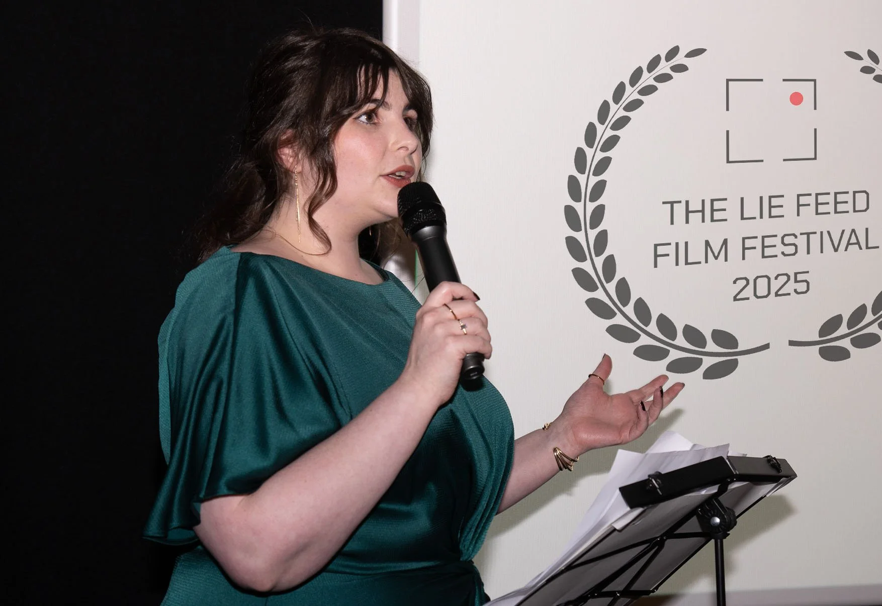 A woman in a teal dress holding a microphone and speaking at The Lie Feed Film Festival 2025, with a white wall displaying the festival's logo and text.