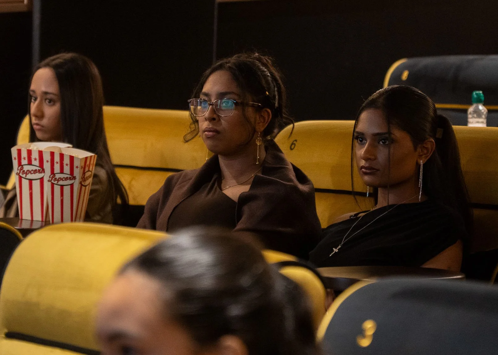 Three women seated in a movie theater, watching a film. The woman in the center is wearing glasses and has dreadlocks, the woman on the right has long dark hair and earrings, and the woman on the left has straight dark hair and a popcorn box in front