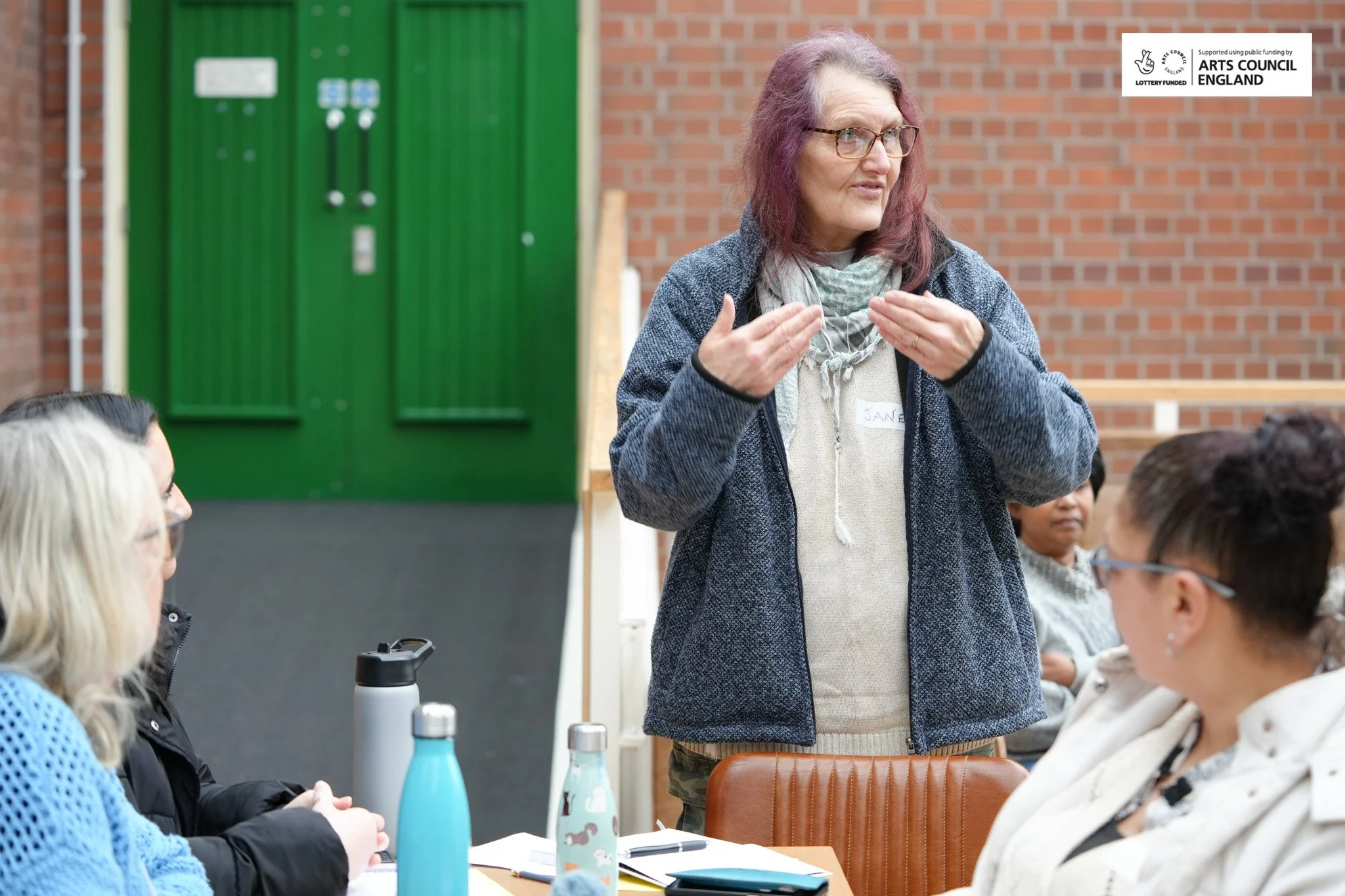 A woman with purple hair and glasses speaking to a group of women in a discussion or presentation, seated at a table with notebooks, pens, and water bottles, in an indoor setting with brick walls and a green door.