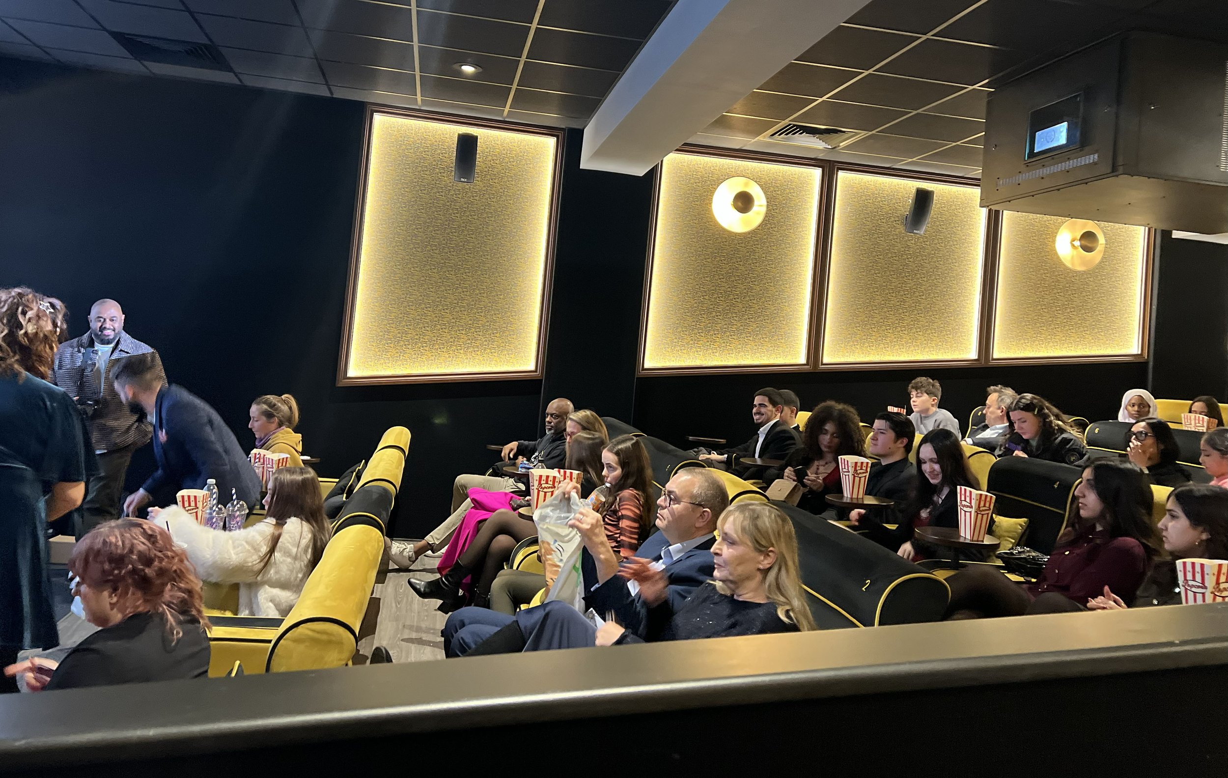 People seated in a darkened theater with yellow and black seats, some enjoying popcorn, others clapping or using their phones, and a few standing near the front.