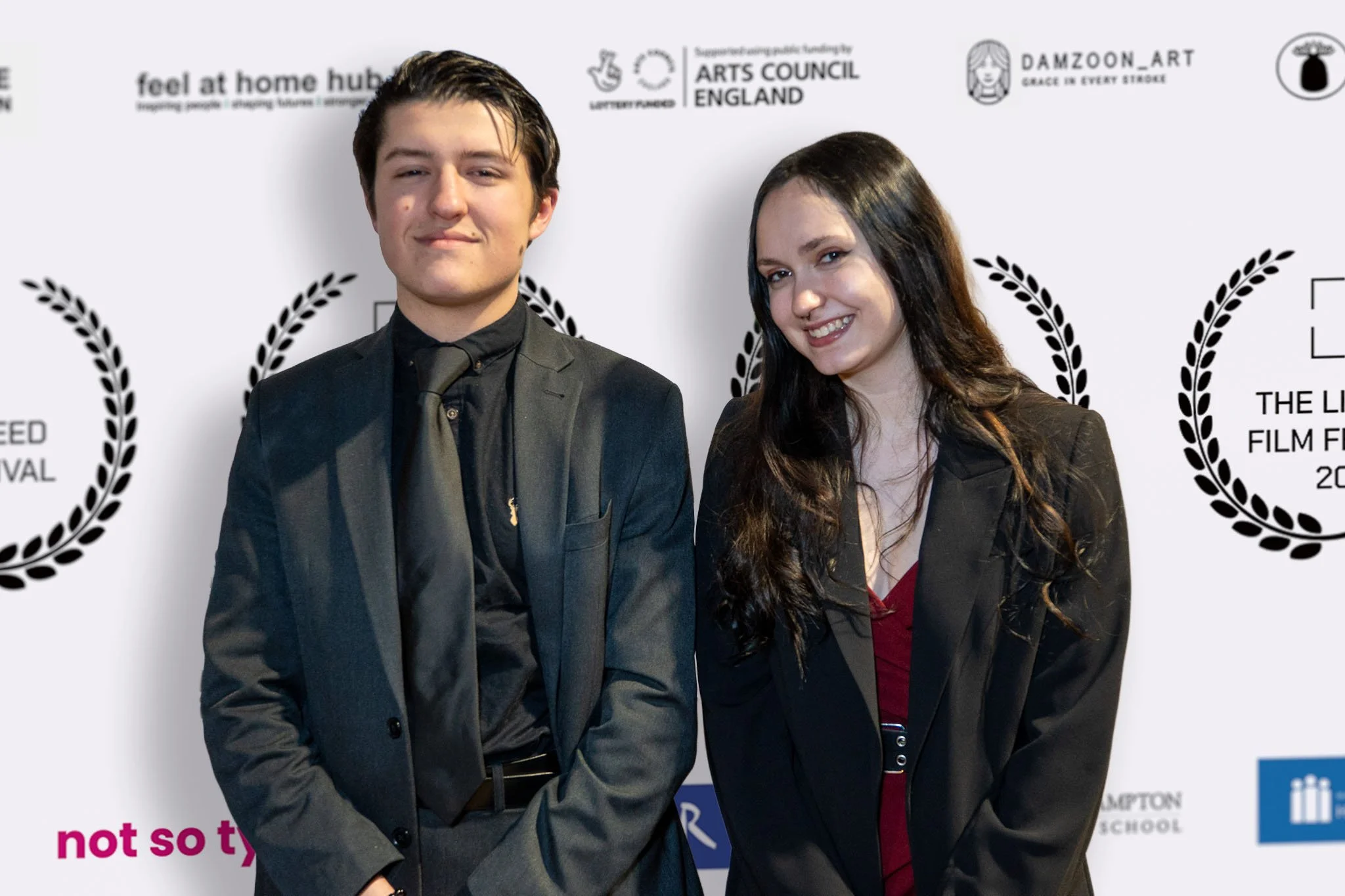 Two young adults, a man and a woman, dressed in formal black attire, standing in front of a backdrop with logos and laurel wreaths at a film festival event.