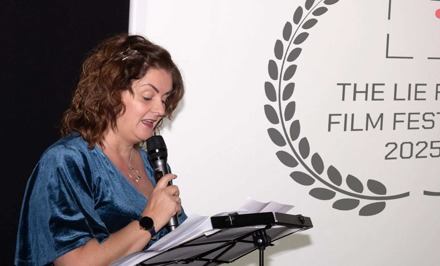 Woman with brown curly hair wearing a shiny blue dress speaking into a microphone while reading from papers at an event. In the background, a sign reads 'THE LIE F ILM FES 2025' within a laurel wreath design.