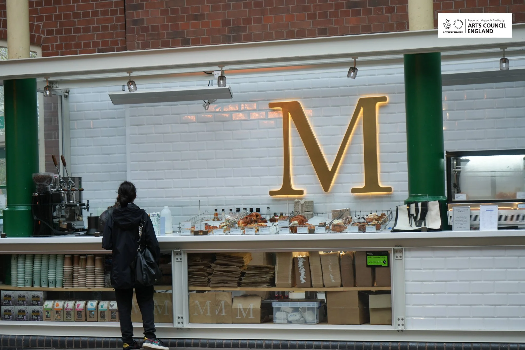 A person stands at a food stall with a prominent illuminated letter "M" on a white brick wall behind it, offering pastries and beverages, with supplies and boxes underneath.